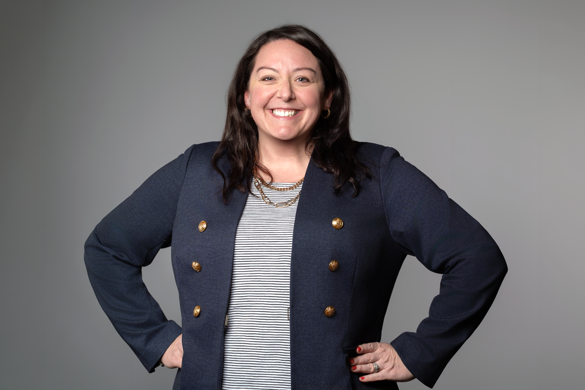 Heather Siefer, promoted at encompas from Project Lead to Market Director in 2026. White woman with dark brown wavy hair standing with hands on hips, smiling and wearing a striped shirt with a navy blue blazer.