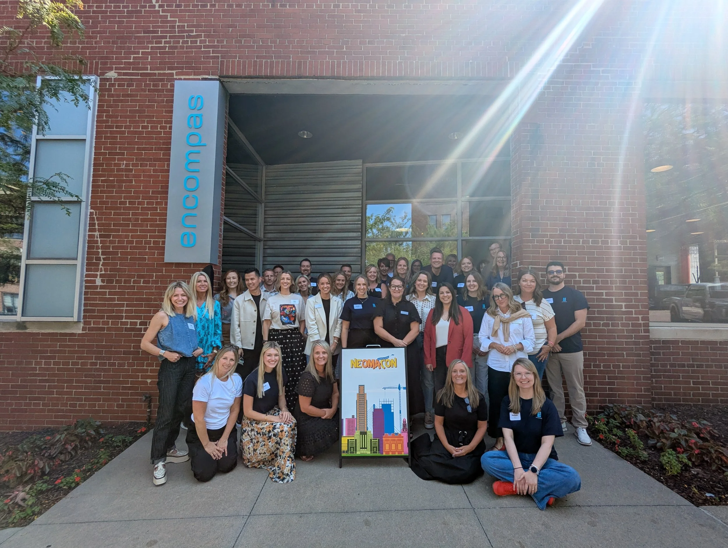 Group photo of people standing and sitting in front of encompas brick building behind sign that says "NeOMAcon"