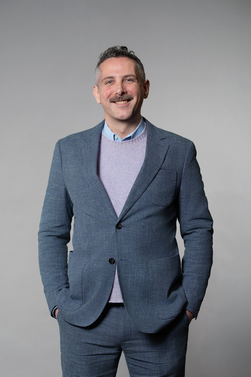 Ian Crawford, promoted at encompas from Project Lead to Market Director in 2026. White man with gray mustache standing confidently and relaxed, smiling and wearing a blue suit with a lavender sweater.