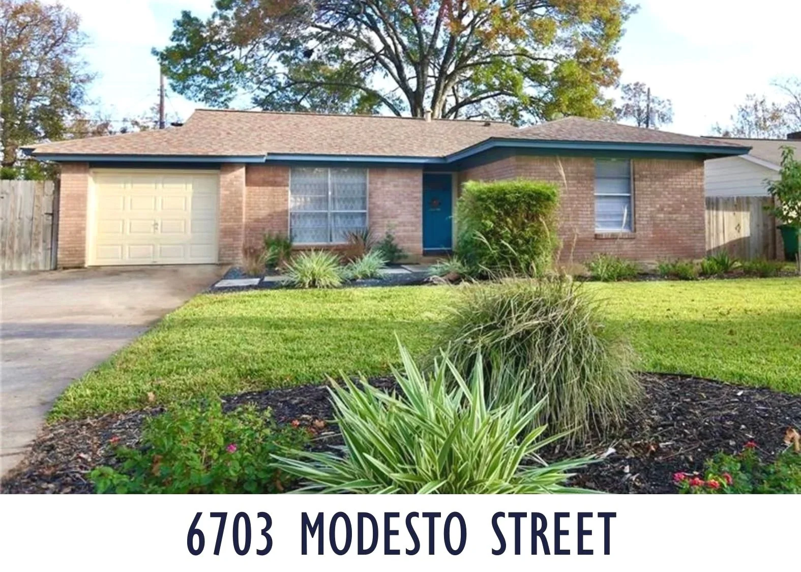 Front view of a single-story brick house with a small front yard, landscaped with green grass, bushes, and plants. The house has a driveway leading to a closed garage door, a blue front door, and two windows with blinds. The roof is shingled, and the