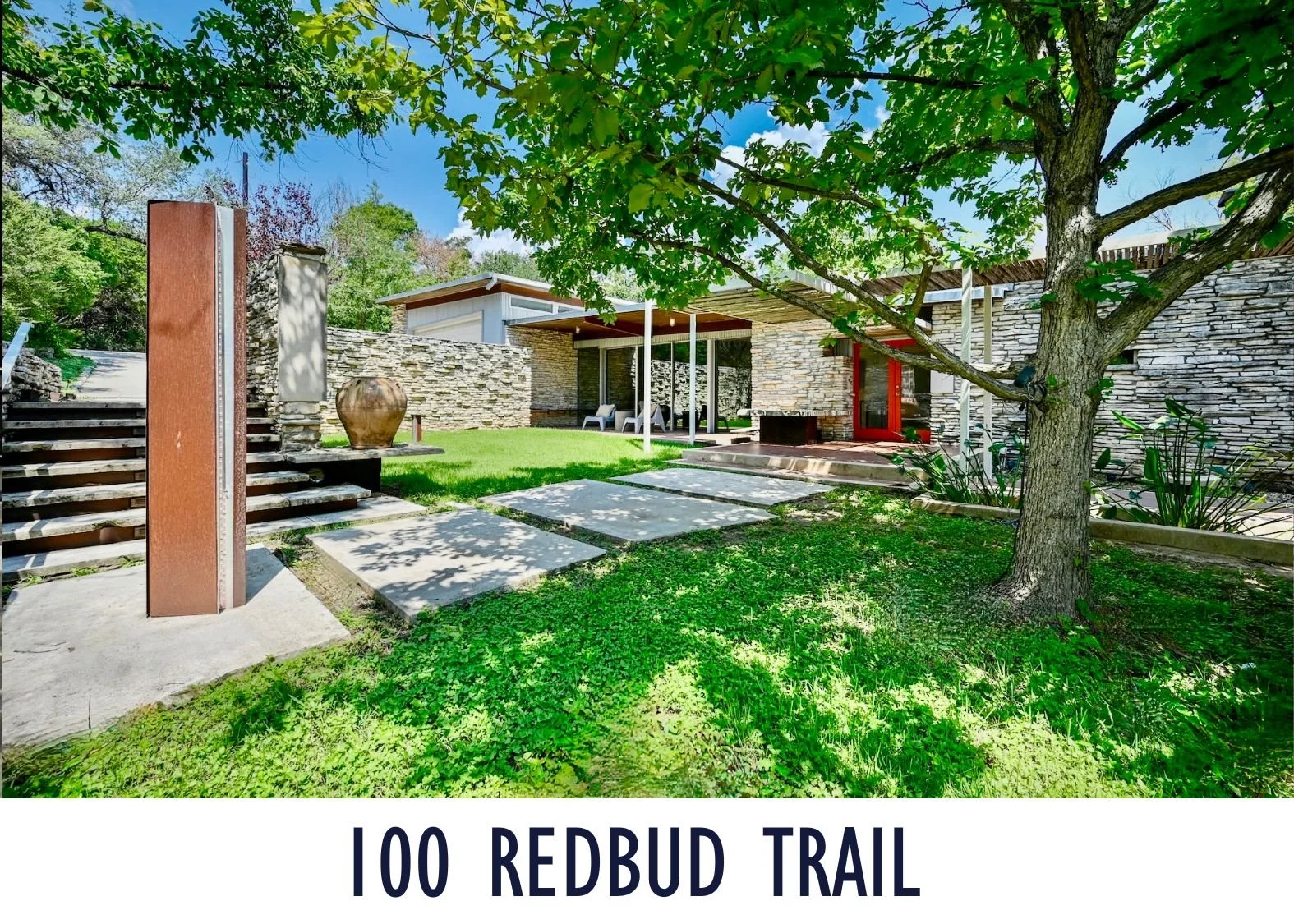 A landscaped backyard with a large tree, stone patio, grass area, and a modern house with large windows and a covered porch. There is a sculpture or water feature to the left and steps leading up to a pathway.