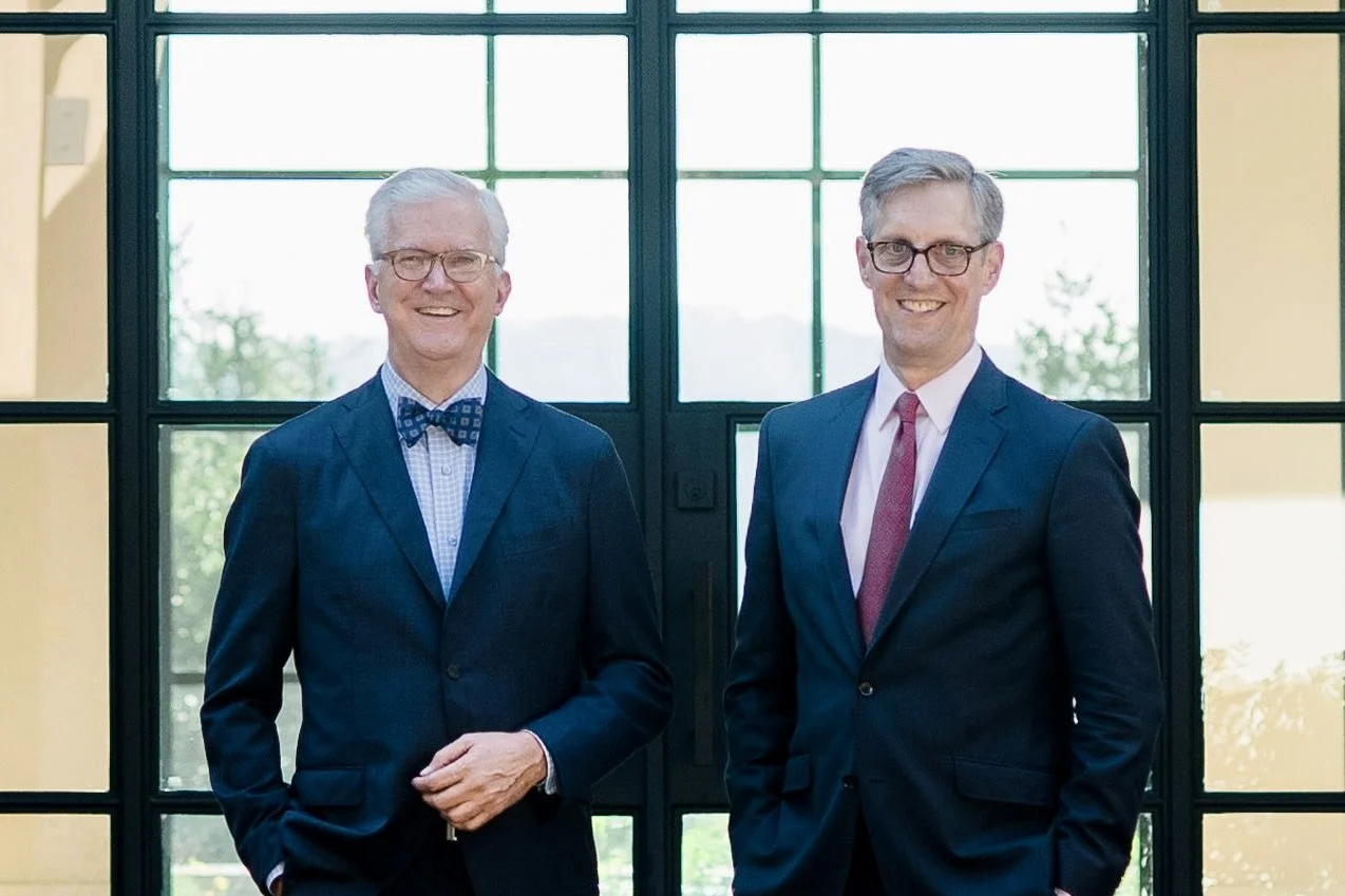 Two men in suits standing indoors in front of large windows, smiling at the camera.