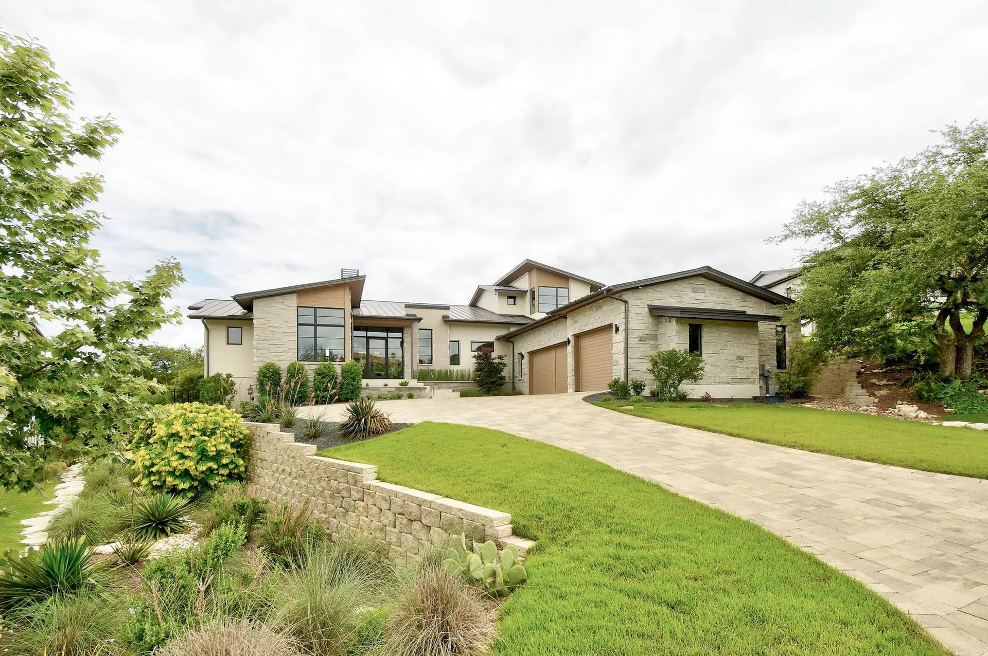 Modern house with large glass windows, stone and stucco exterior, surrounded by landscaped garden and paved driveway.