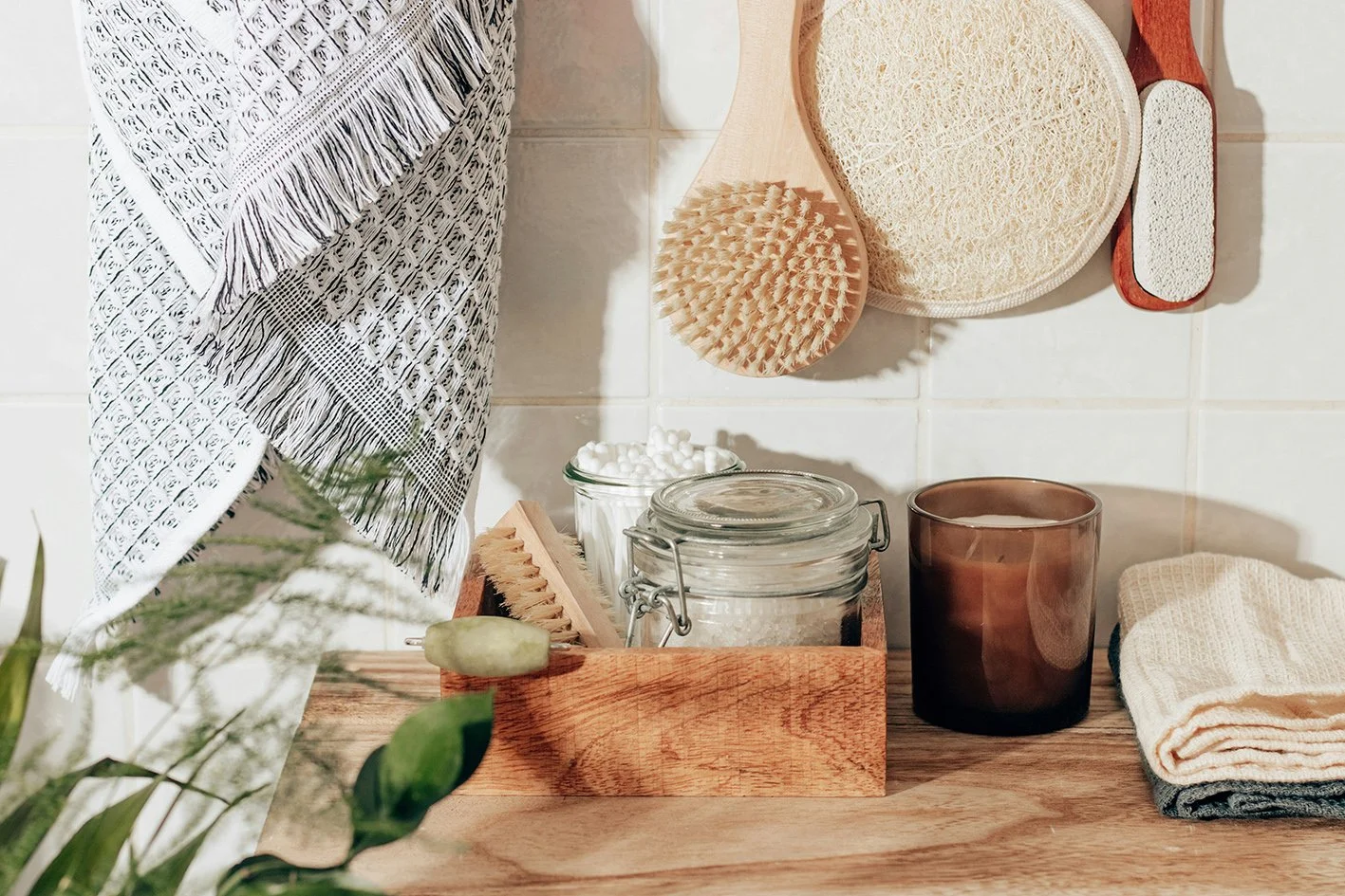 Bathroom shelf with natural skincare tools, a glass jar of cotton swabs, a brown candle holder, and folded towels on a wooden surface; hanging on the wall are bath brushes and scrubbers.