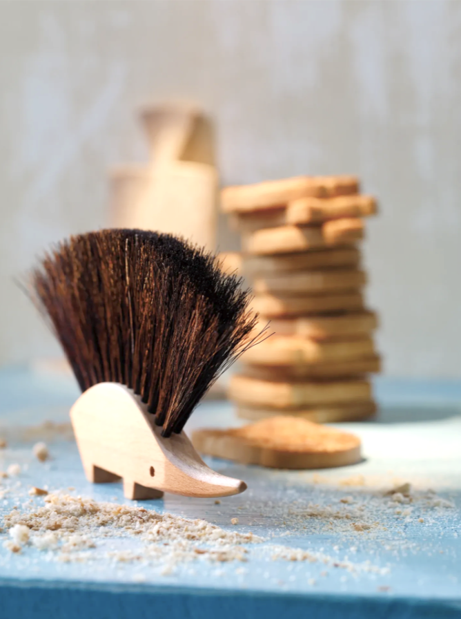 A small wooden hedgehog brush with coarse bristles on a blue surface, with scattered wood shavings and a blurred background of stacked wooden pieces and a white object.