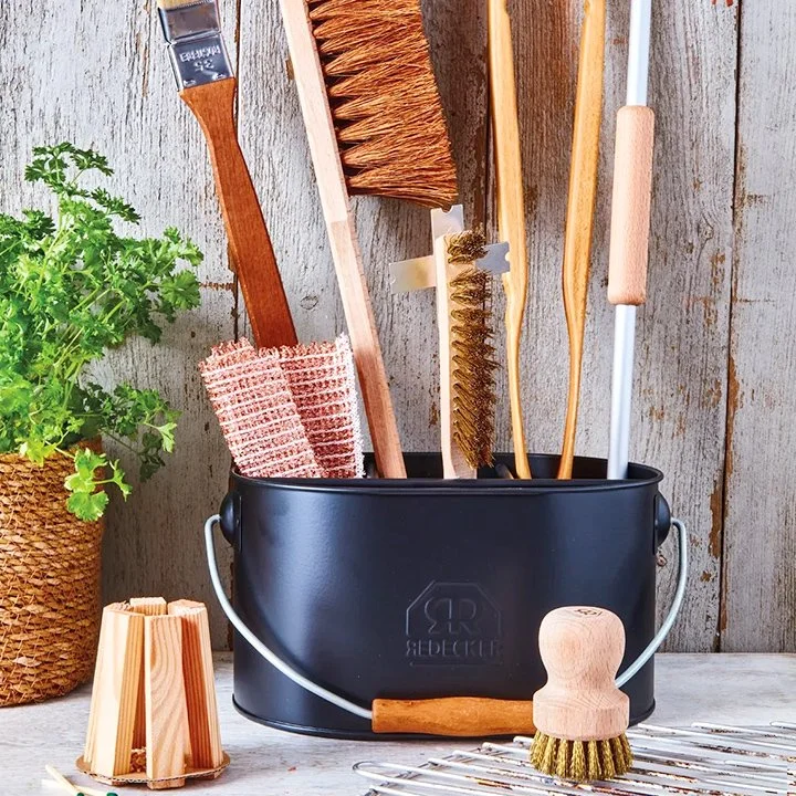 A collection of cleaning brushes and tools in a black container on a rustic wooden surface, next to a potted green plant.