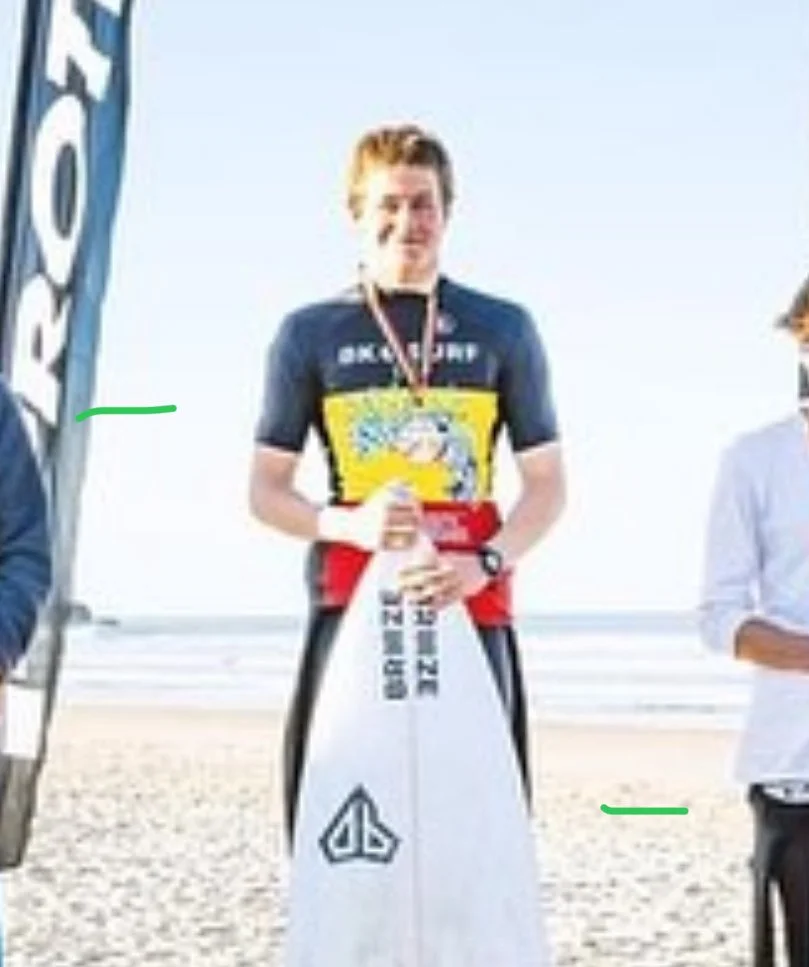 Winner's podium at a surfing competition with three young male surfers standing on the podium, medals around their necks, and one holding a microphone. Surfboards and surfing equipment are visible, with a DJ or announcer in the background.