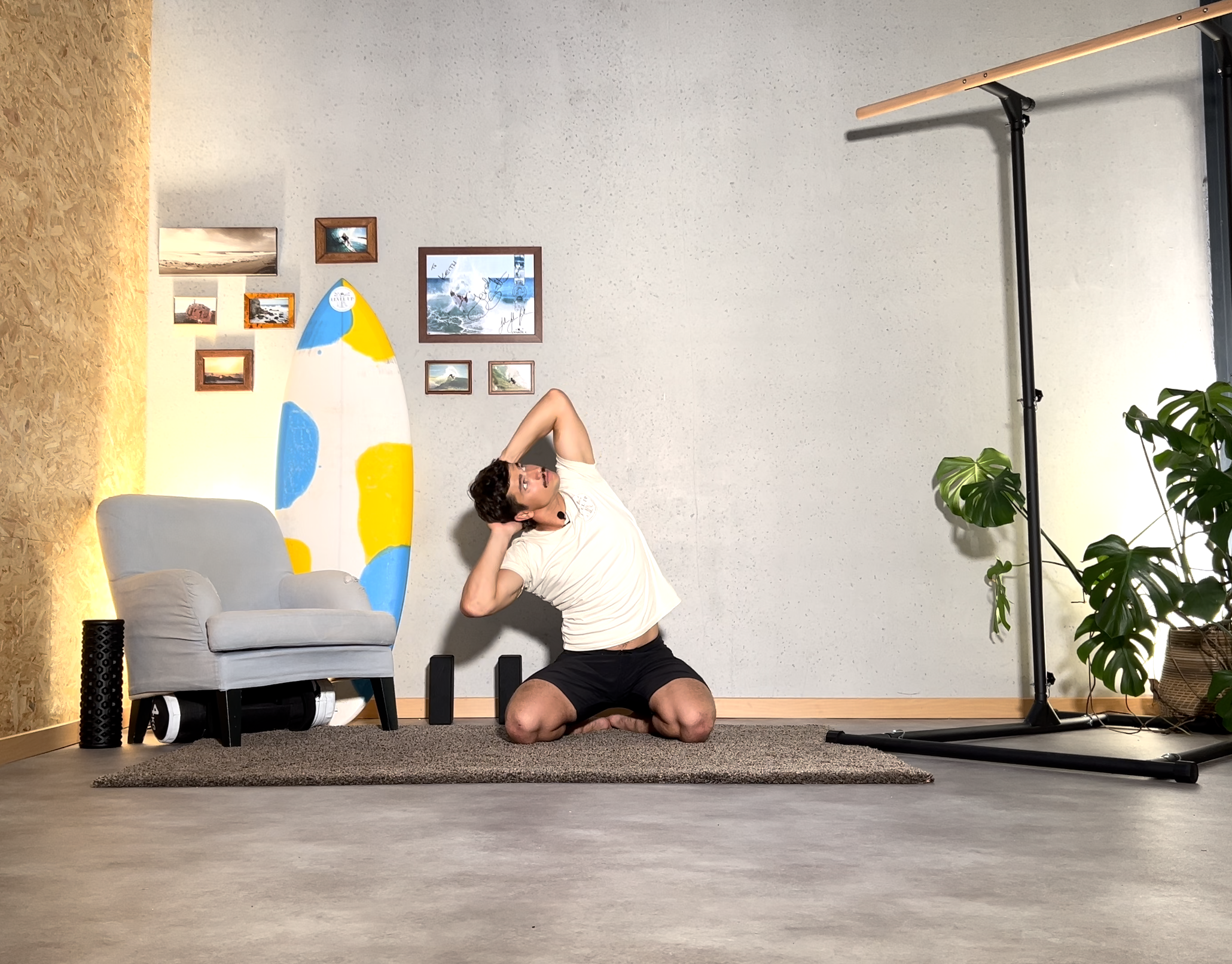 A man doing a stretch exercise on his knees in a room with a surfboard leaning against the wall, a small couch, a picture of a surfboard, and a large potted plant.