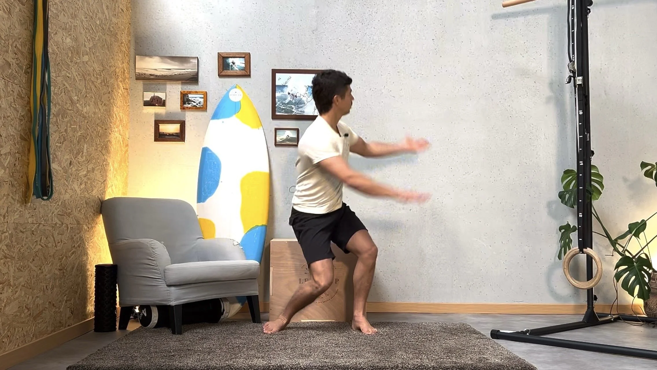 A young man in a white shirt and black shorts appears to be doing a workout or exercise indoors in a living room. The room has a gray armchair, a surfboard leaning against the wall with a blue, yellow, and white design, and several framed pictures hanging on the wall. There are also workout equipment including a ring and a PVC pipe, and a potted plant.