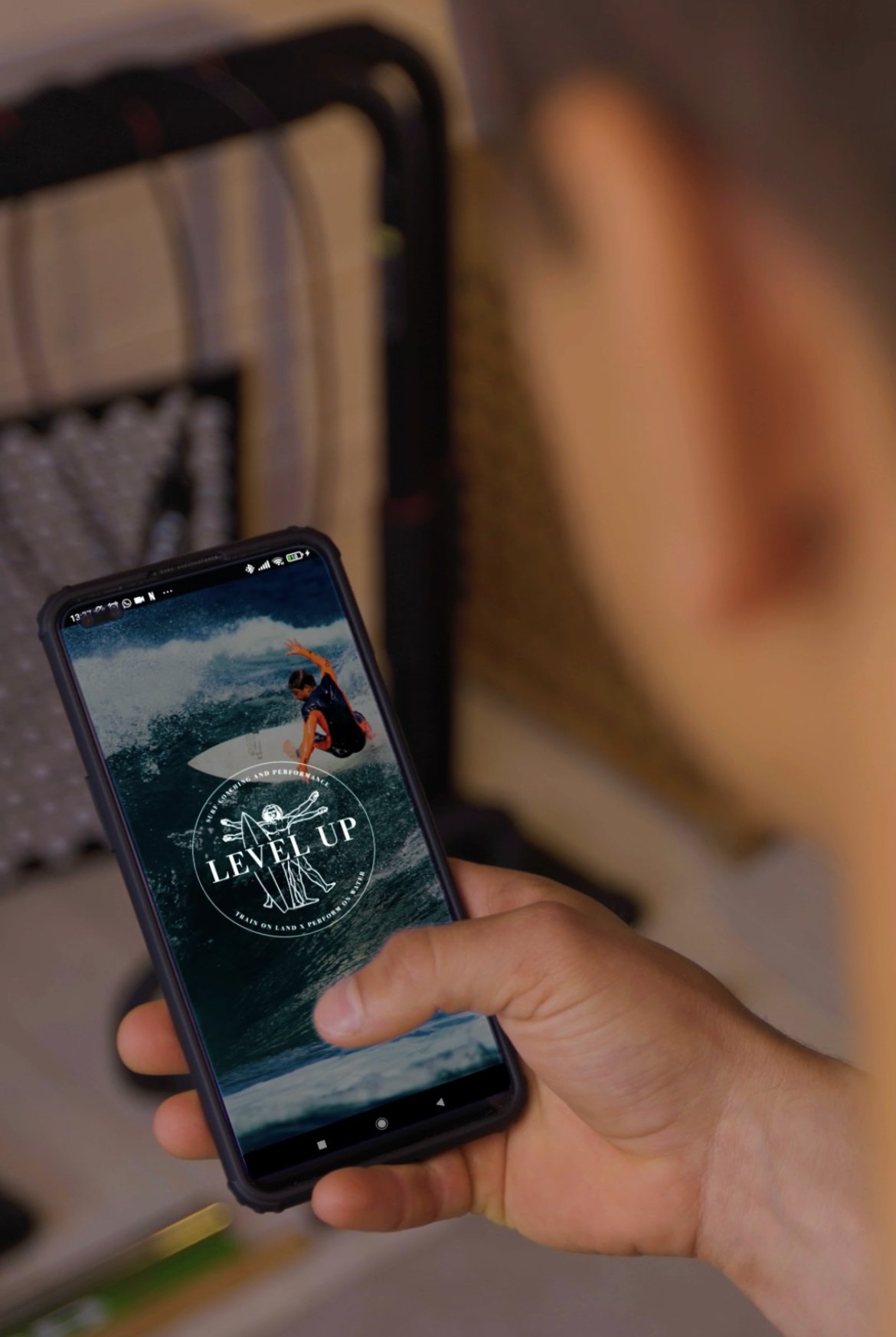 A hand holding a smartphone displaying a surfing app with an image of a surfer riding a wave and the text 'LEVEL UP'.