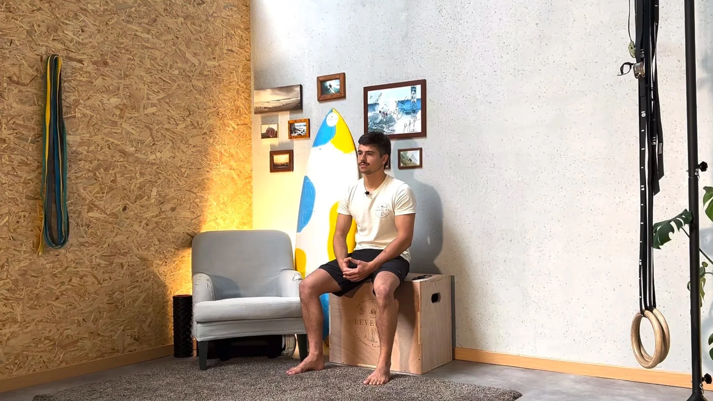 A man is sitting on a wooden box in a room with motivational decor, a white armchair, and a surfboard wall art. There are framed pictures on the wall, and gym rings and resistance bands are visible. The man is barefoot, wearing a white t-shirt and black shorts, and appears to be preparing for fitness training.