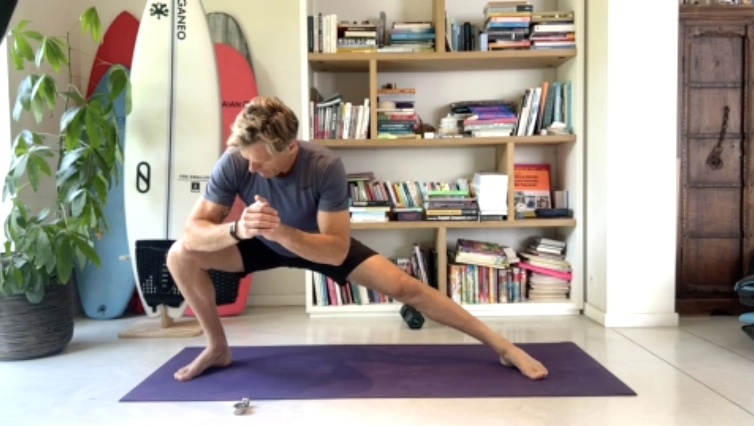 Man practicing yoga in a kneeling split position on a purple yoga mat indoors, with surfboards and a bookshelf in the background.