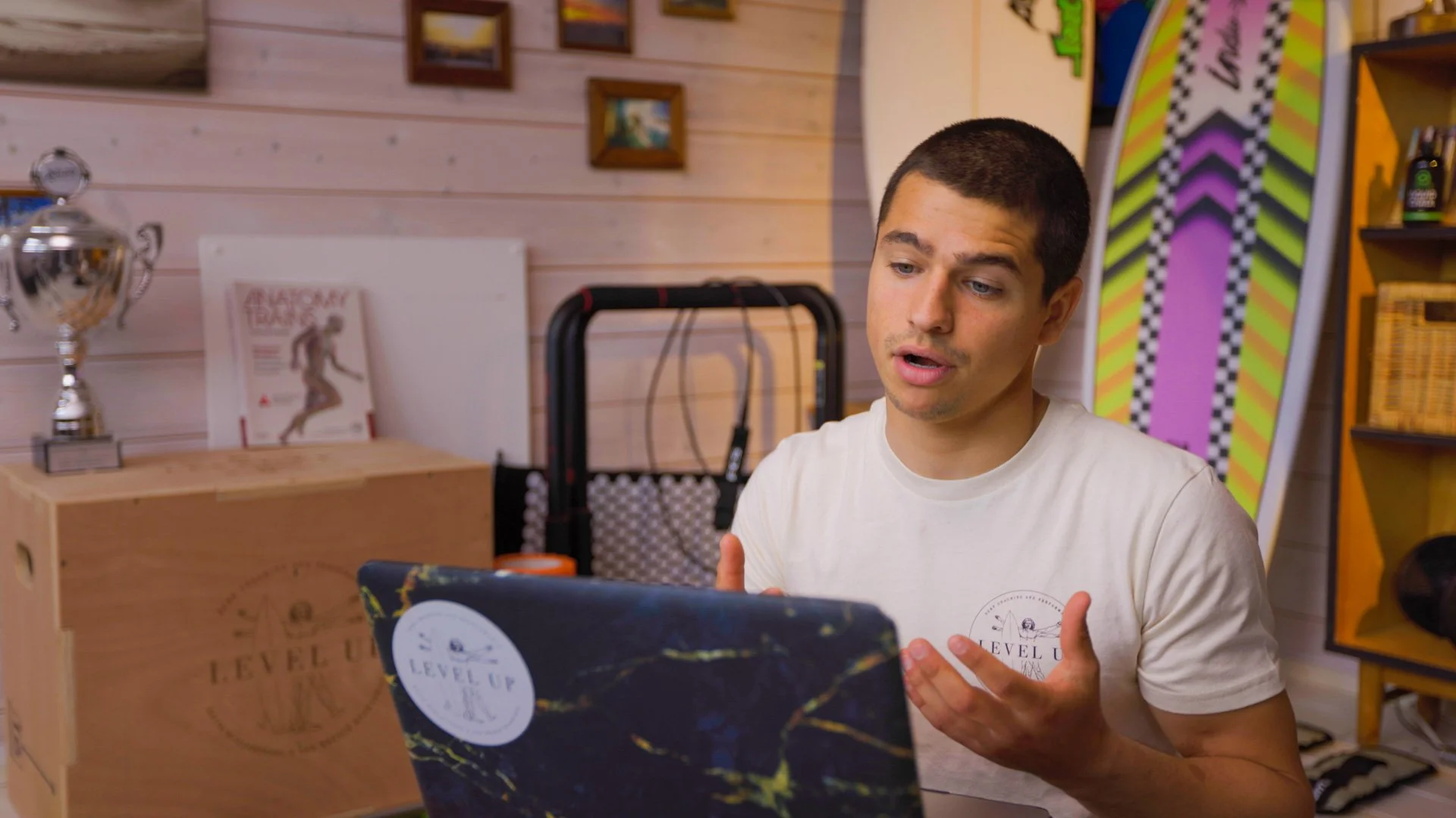 Young man with short dark hair and a light beard, wearing a white T-shirt, sitting indoors, engaging with a laptop or tablet, with a wooden wall, colorful artwork, and shelves in the background.
