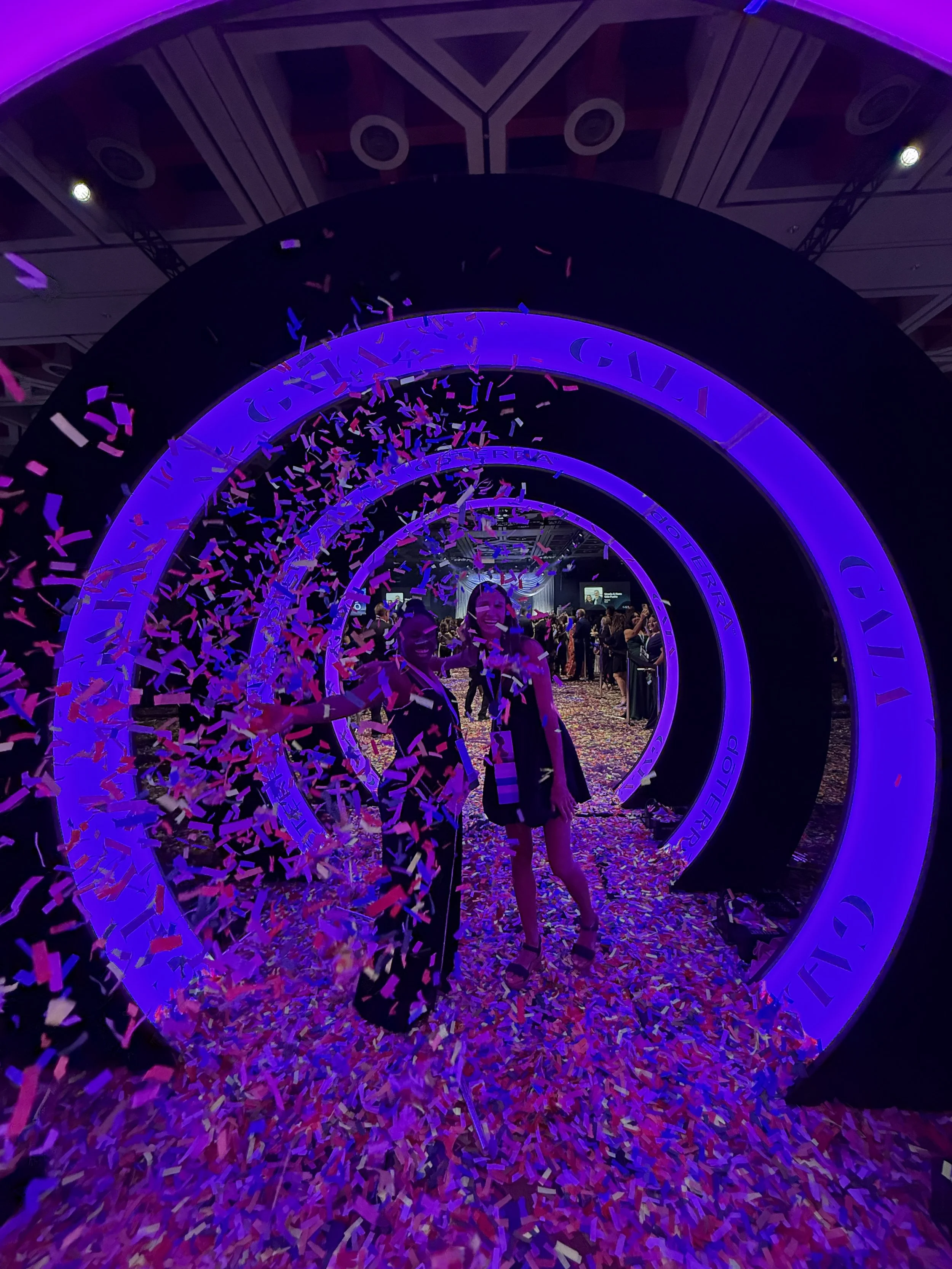 Two women celebrate at a conference with purple neon lighting, standing through circular frames, with colorful confetti falling around them and on the floor, and a crowd of people in the background.