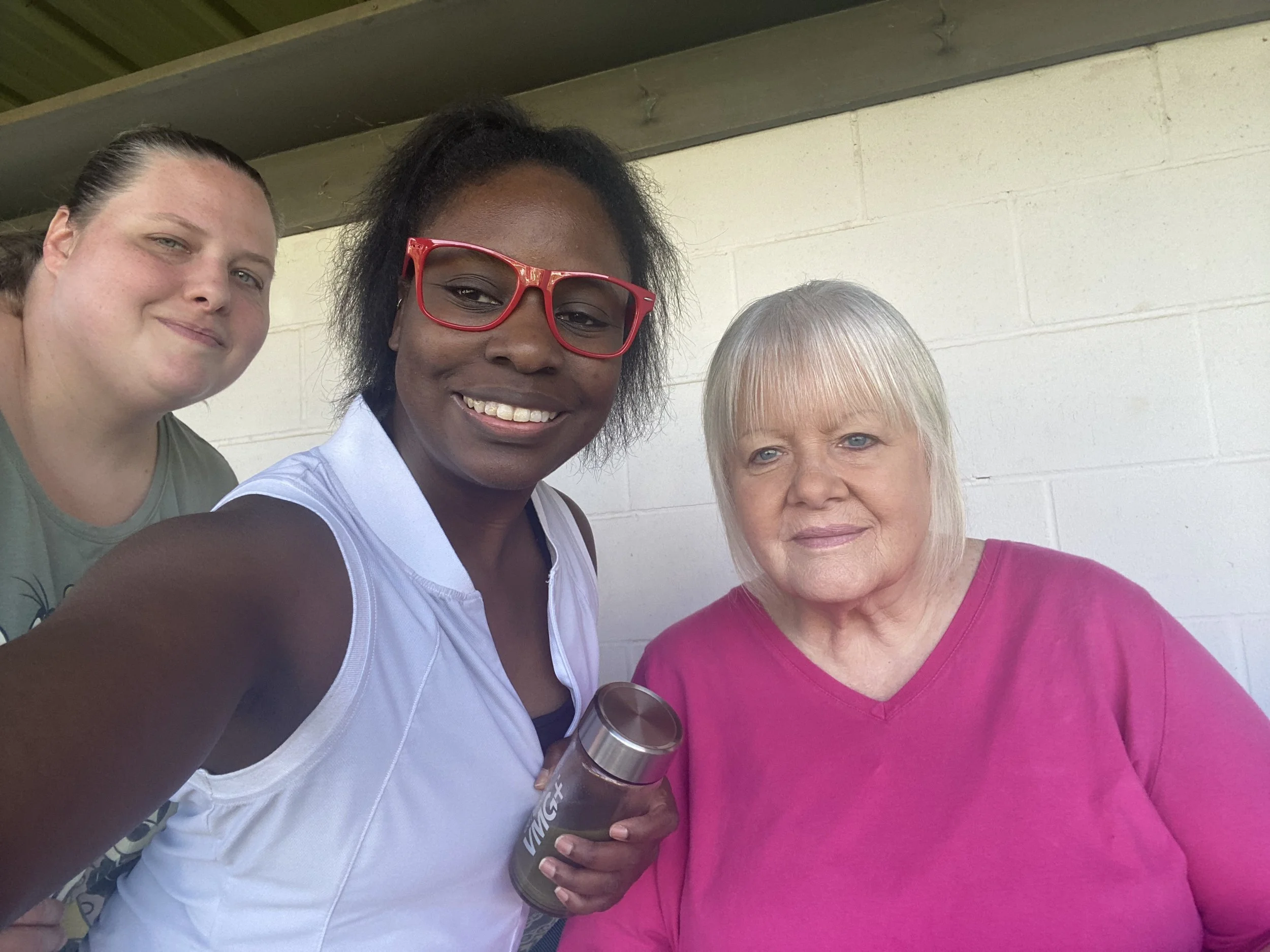 Three women smiling outdoors in front of a light-colored brick wall, one holding a water bottle, one wearing red glasses, and one wearing a pink shirt.