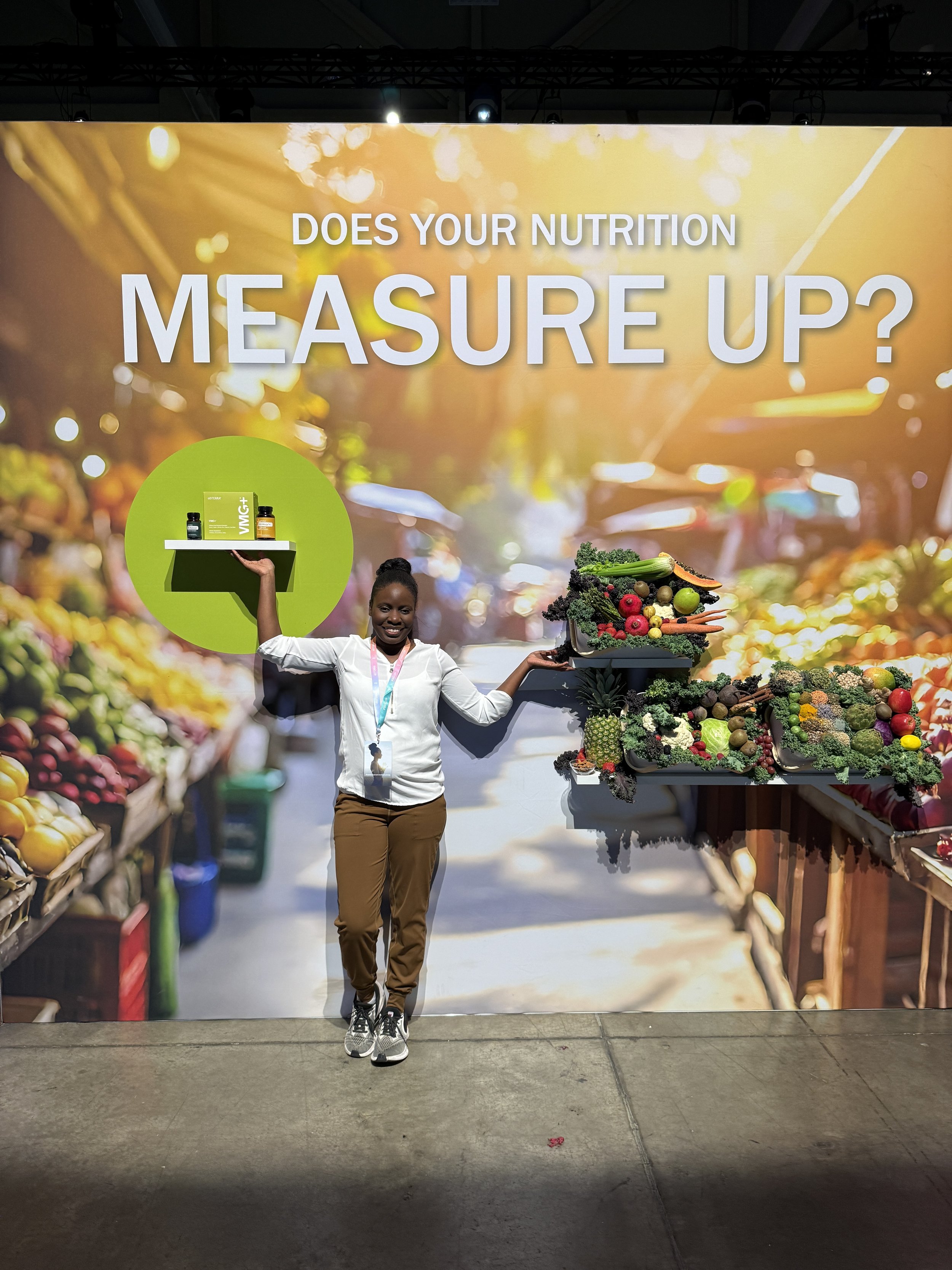A woman at a booth with a large backdrop that reads 'Does your nutrition measure up?' She is smiling, holding scaled tablets with supplement bottles in one hand and a tray of vegetables in the other, at an indoor event or trade show.