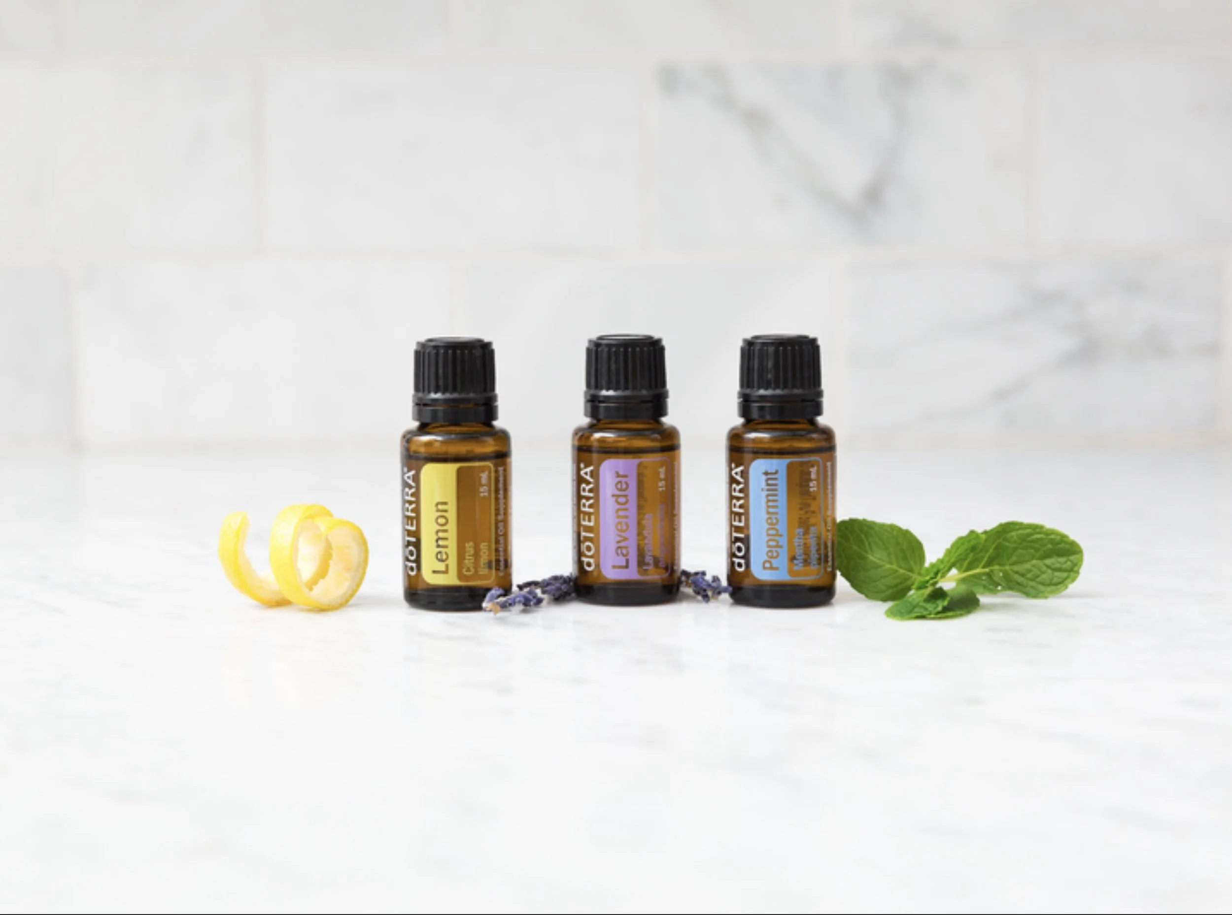 Three bottles of essential oils labeled lemon, lavender, and peppermint on a white surface with lemon peel, lavender flowers, and mint leaves around them.