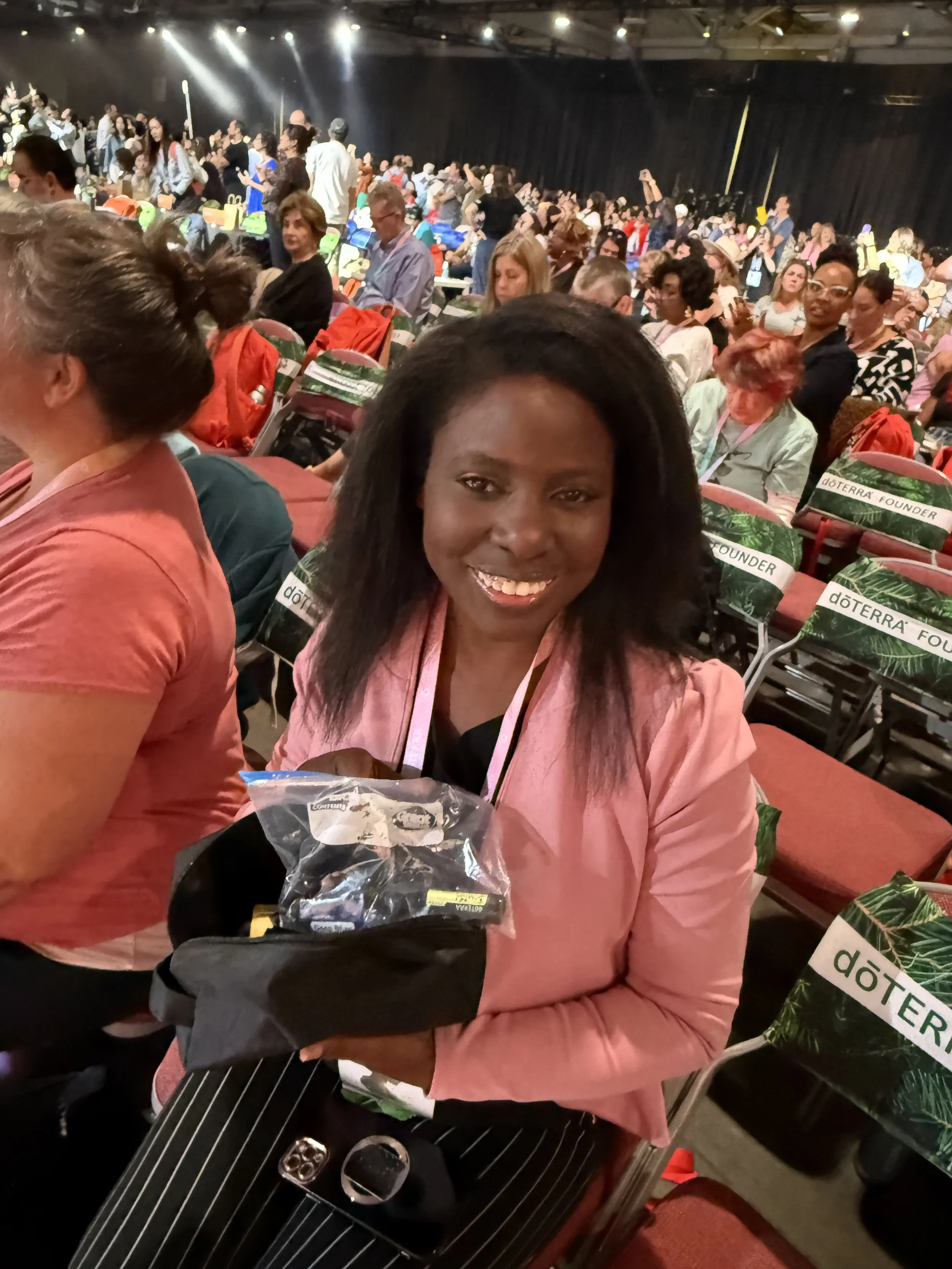 Smiling woman sitting in auditorium holding a gift bag at a conference event with many attendees in the background.