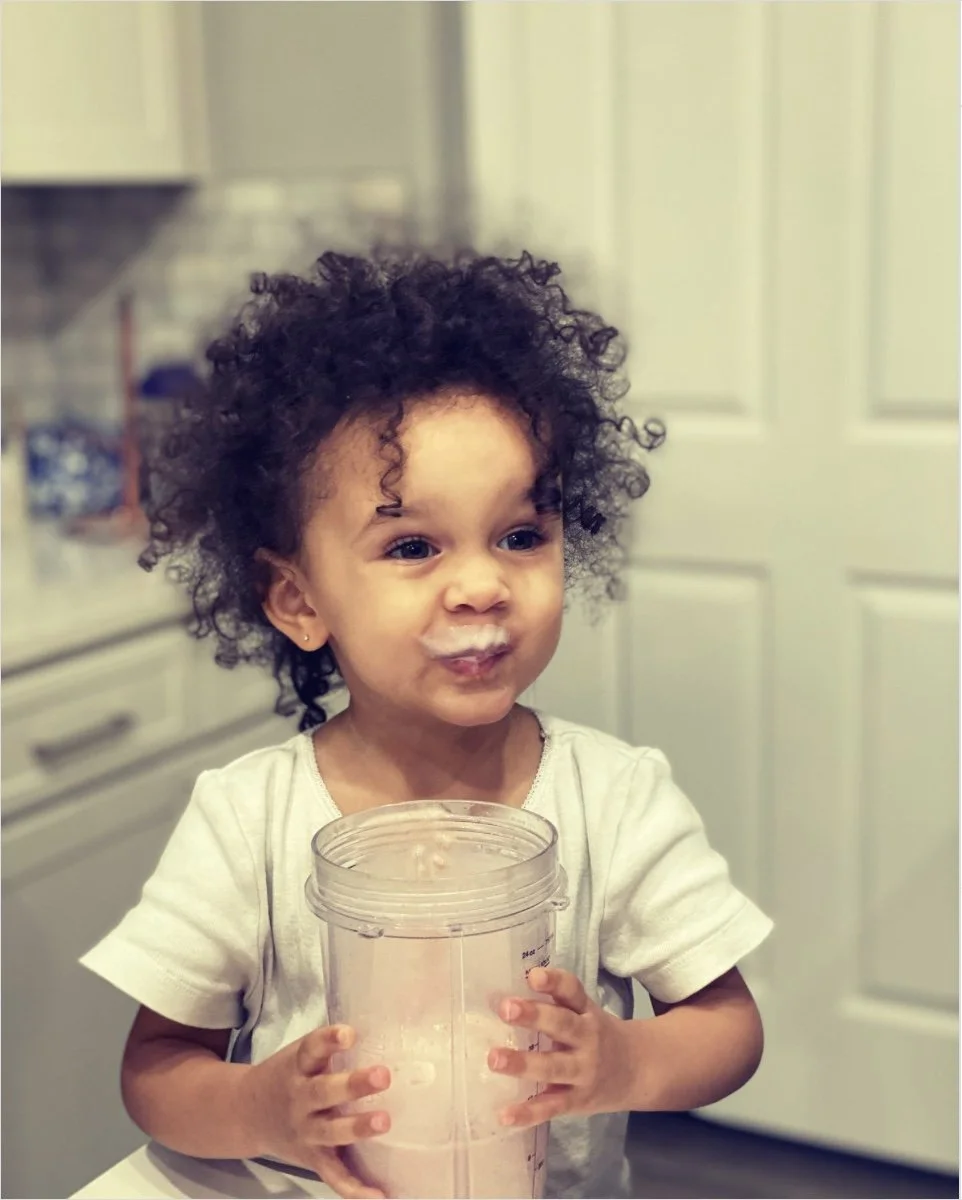 A young girl with curly hair holds an open blender jar, smudged with a white substance around her mouth, in a kitchen with cream-colored cabinets.