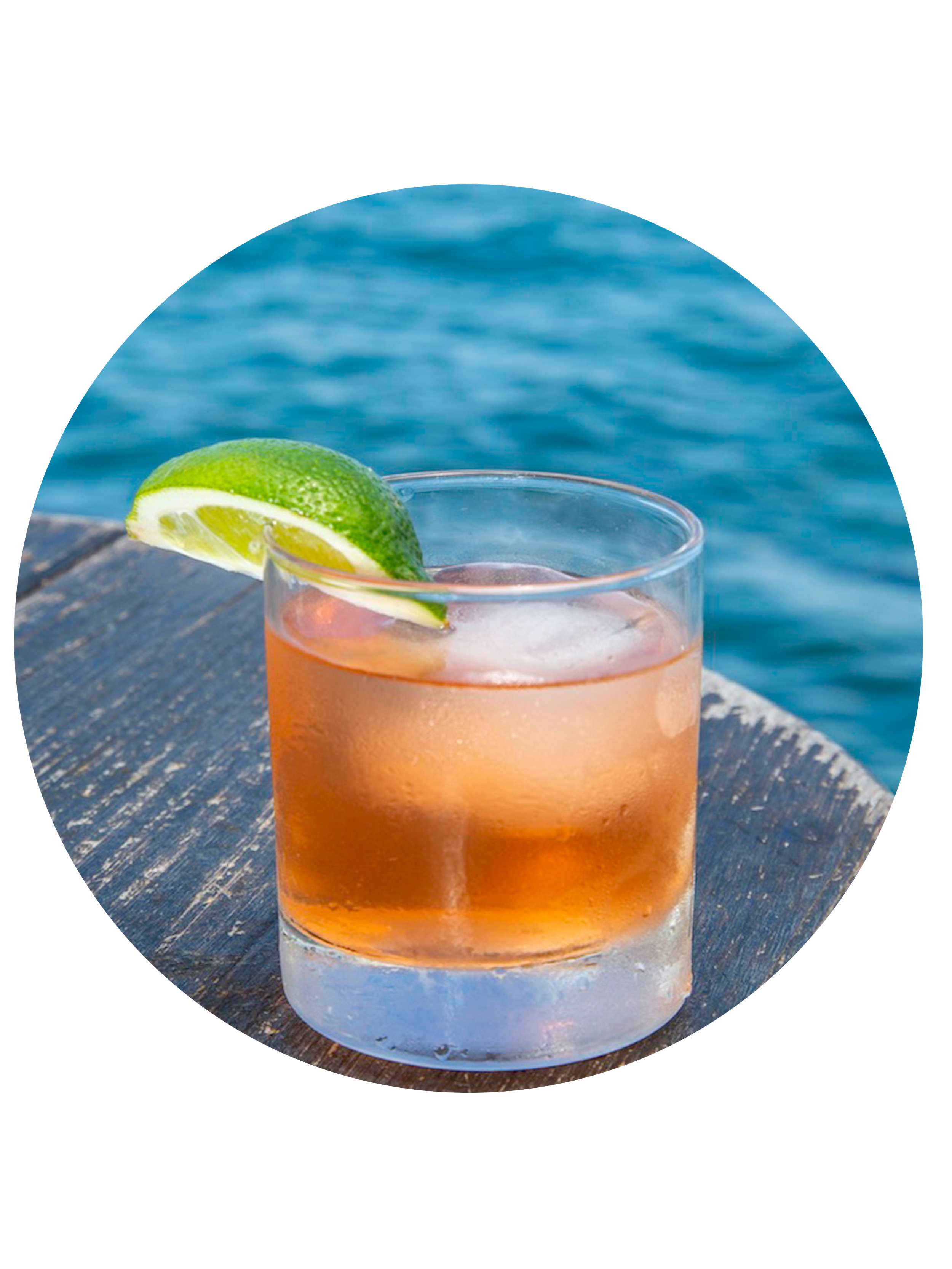 A glass of pink cocktail with ice and a lime wedge garnish on a wooden surface near water.