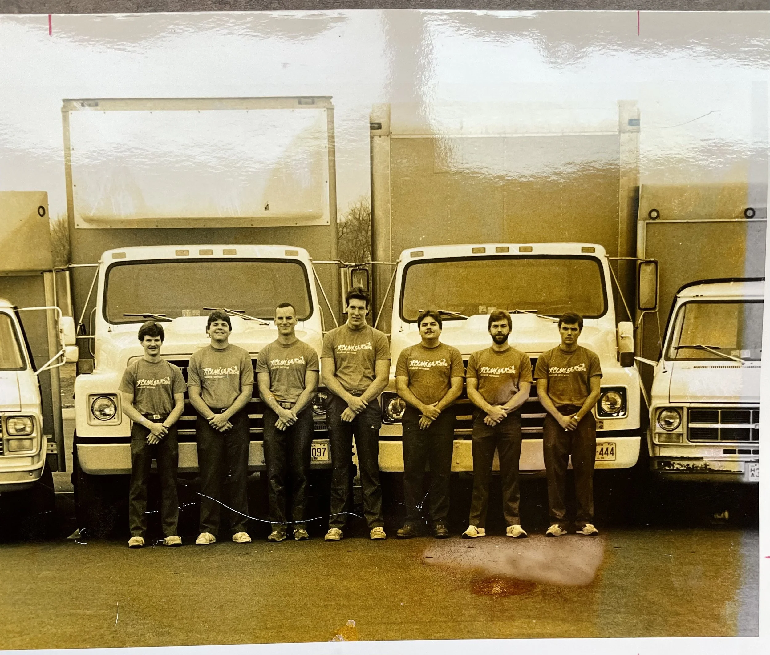 Group of seven people standing in front of three large trucks, all wearing matching T-shirts and standing outdoors.