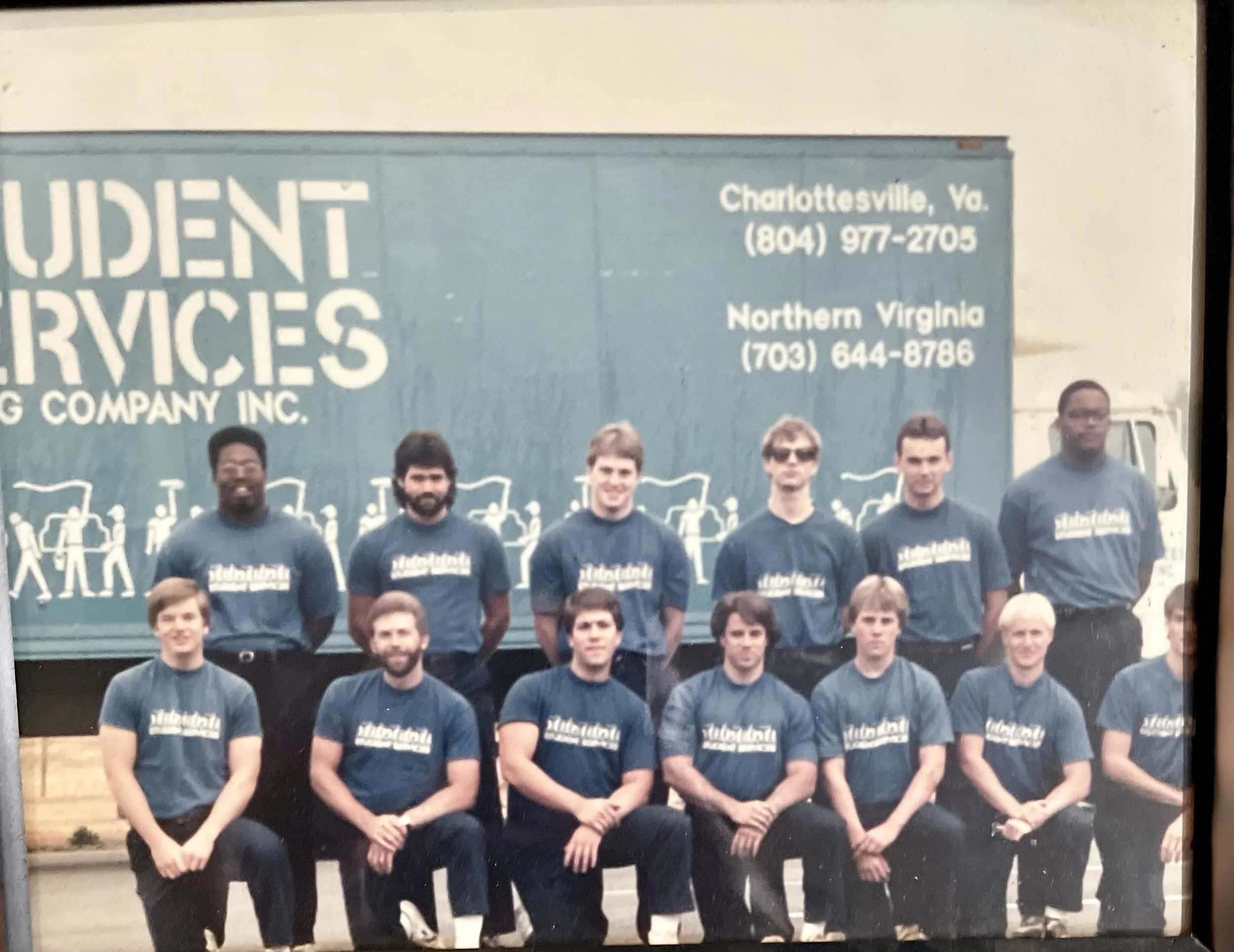 A group of ten young men in matching blue shirts posing in front of a large truck with a logo for Student Services. The truck has contact information for locations in Charlottesville, VA, and Northern Virginia. The men are standing and sitting in two rows outdoors.