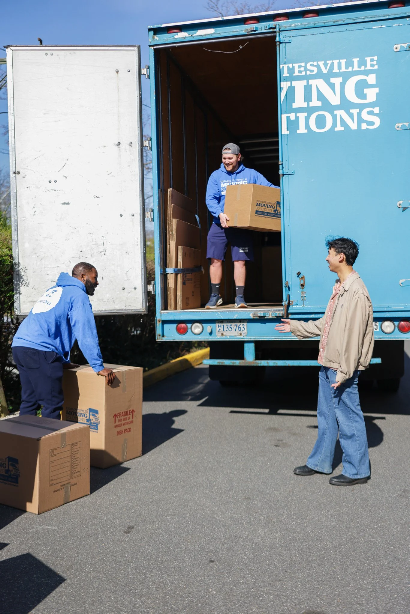 People unloading boxes from a moving truck labeled 'Charlottesville Moving Solutions' in a parking lot on a sunny day.