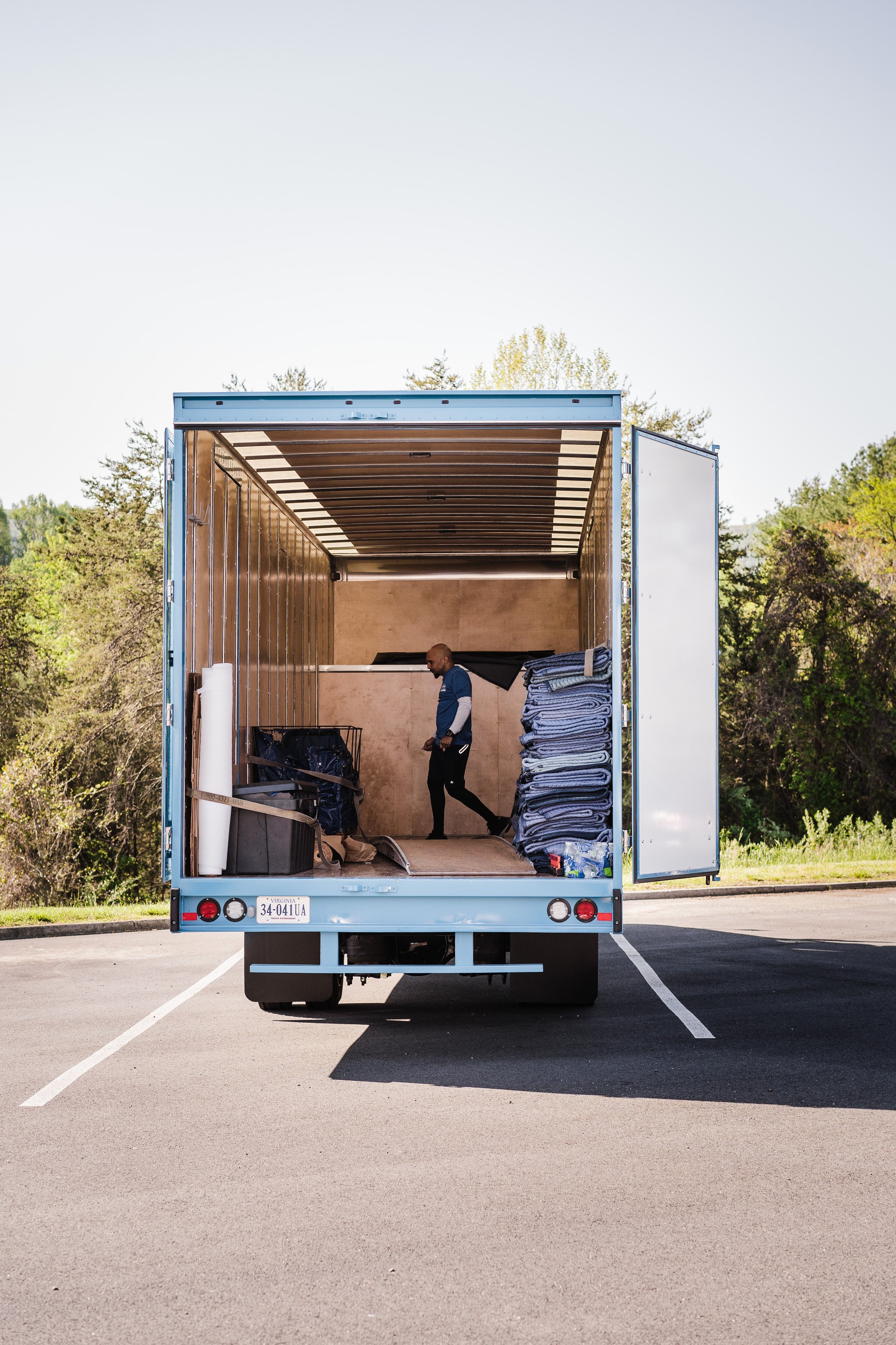 An open moving truck parked in a parking lot with a man inside organizing or moving items, including folded blankets and plastic wrap.