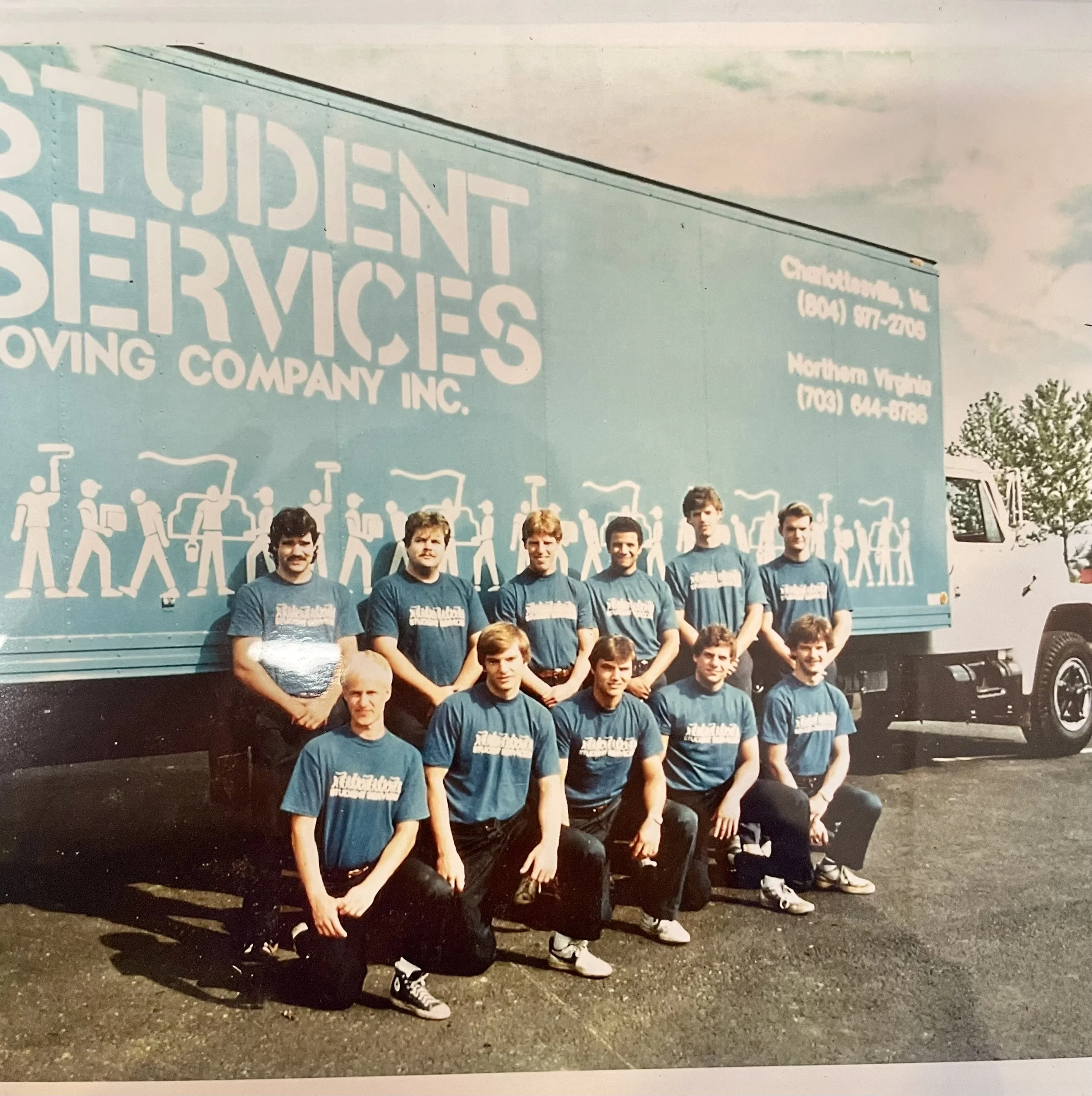 A group of ten young men in matching blue t-shirts with white text, posing in front of a large moving truck with graphics and text on it. The truck advertises a student moving company in Charlotte and Northern Virginia.