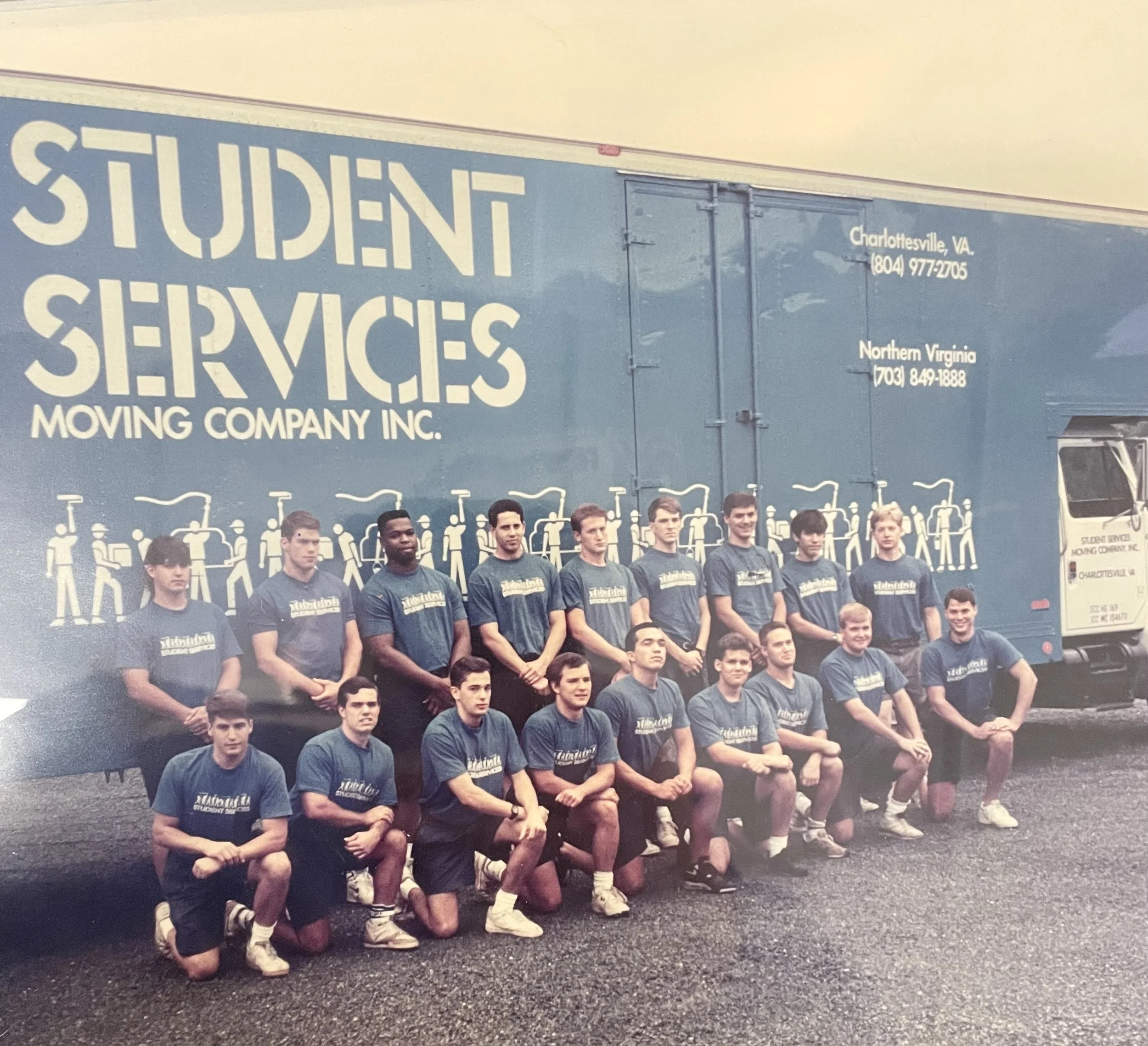 A group of young men in athletic uniforms posing in front of a large moving company's truck that reads 'Student Services Moving Company Inc.' with contact information for Charlottesville, VA, and Northern Virginia.