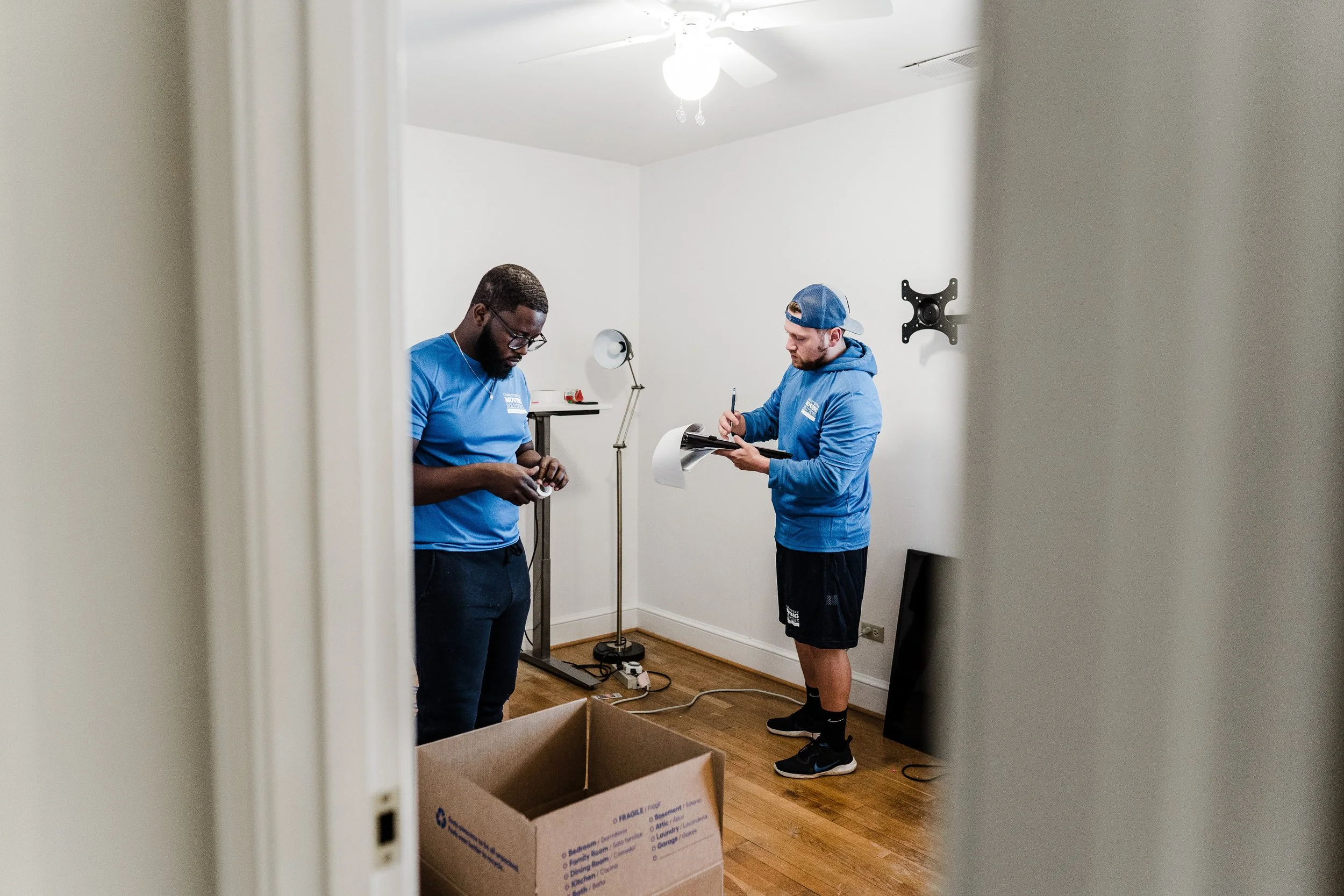 Two men in blue shirts are standing in a room with white walls and a ceiling fan. One man is holding a roll of tape, and the other is taking notes on a clipboard. There are cardboard boxes on the floor and a cardboard box in the foreground. The scene appears to be of a home or office moving or organizing event.