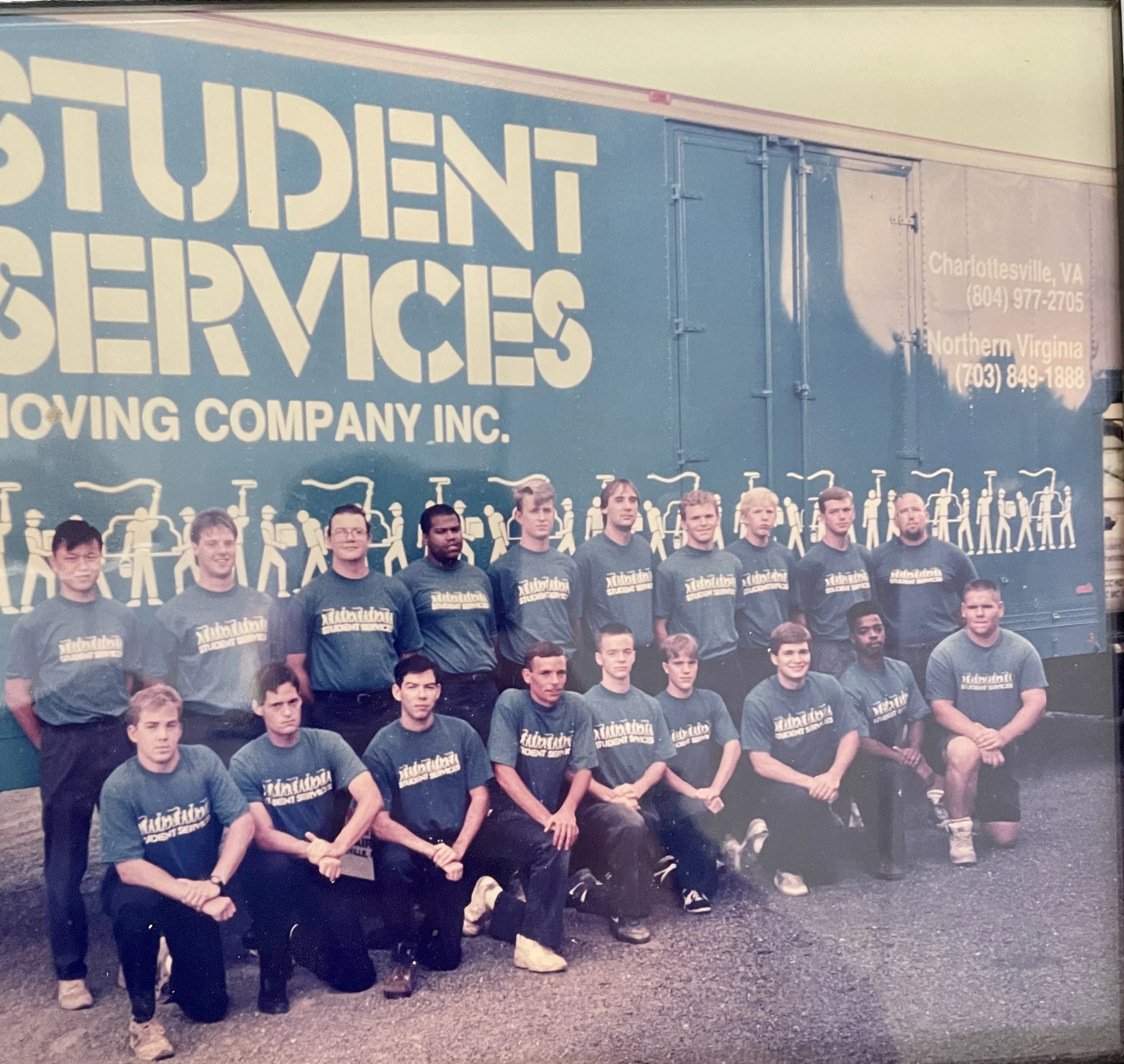 Group of students and staff members in front of a moving services truck, wearing matching T-shirts, posing for a team photo.