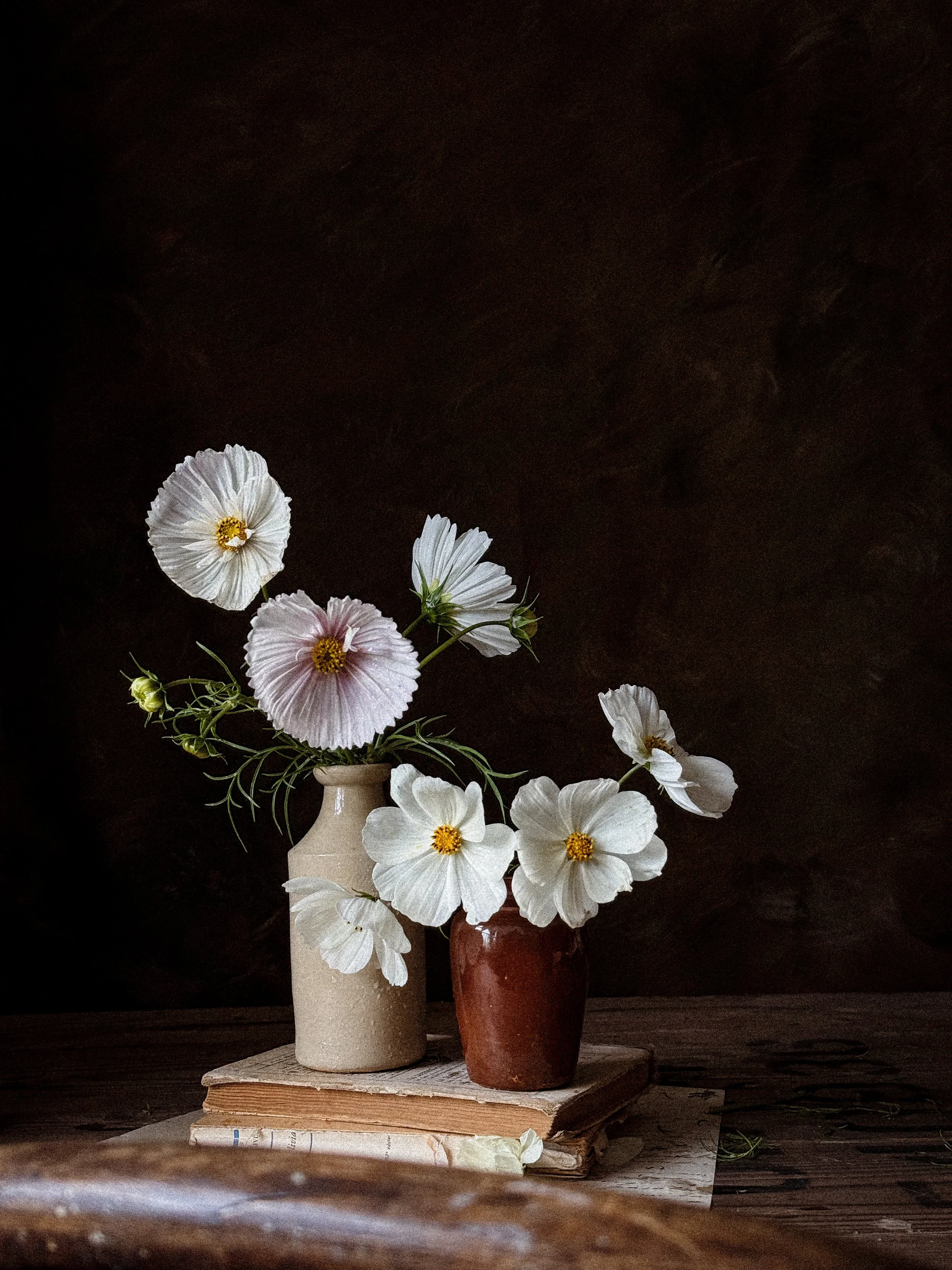 A still life composition featuring white and pink flowers in two ceramic vases, set on a wooden platform with weathered books underneath, against a dark background.