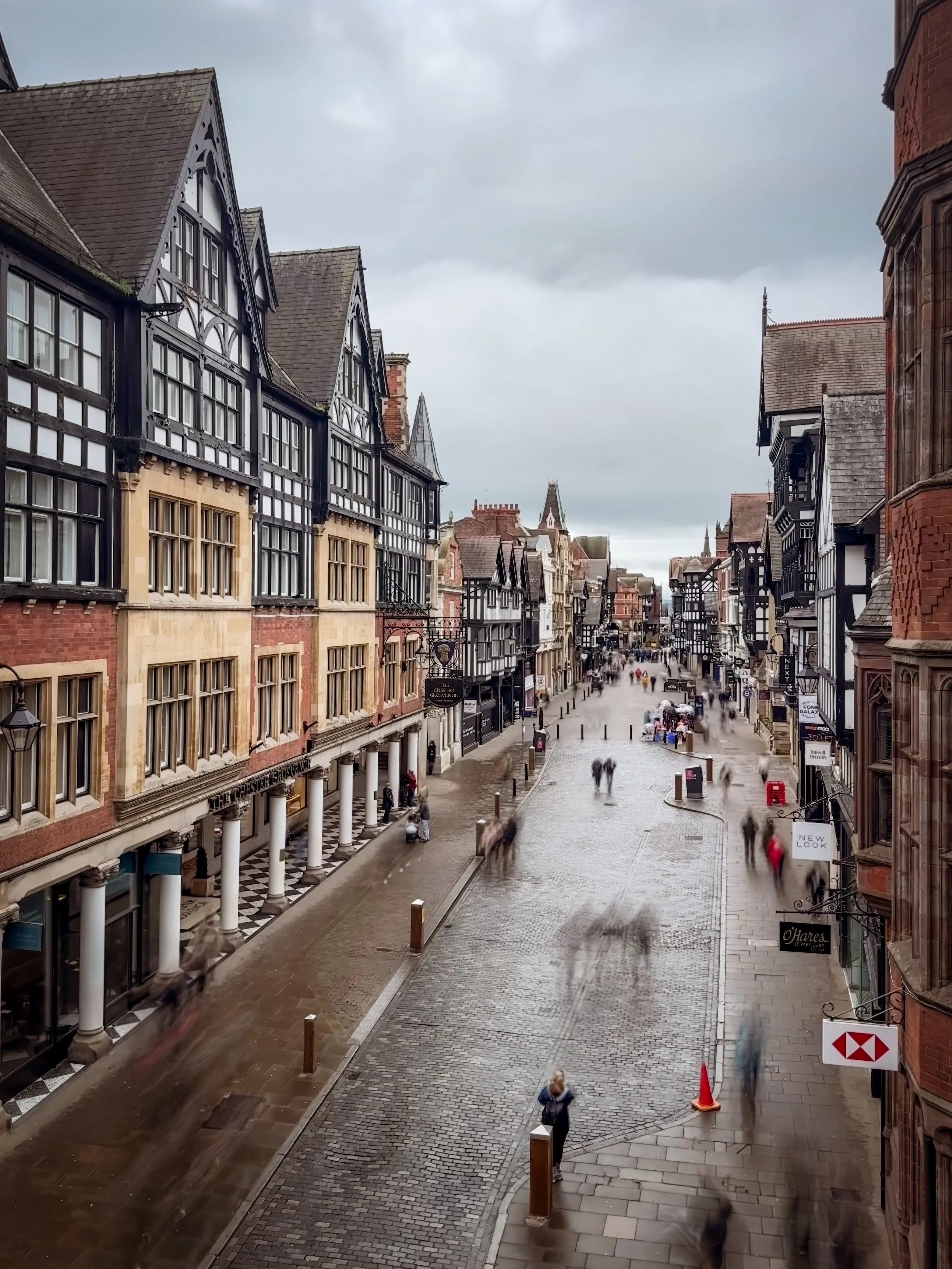 A city street with historic black and white timber-framed buildings on either side, pedestrians walking on wet cobblestone pavement, and cloudy skies overhead.