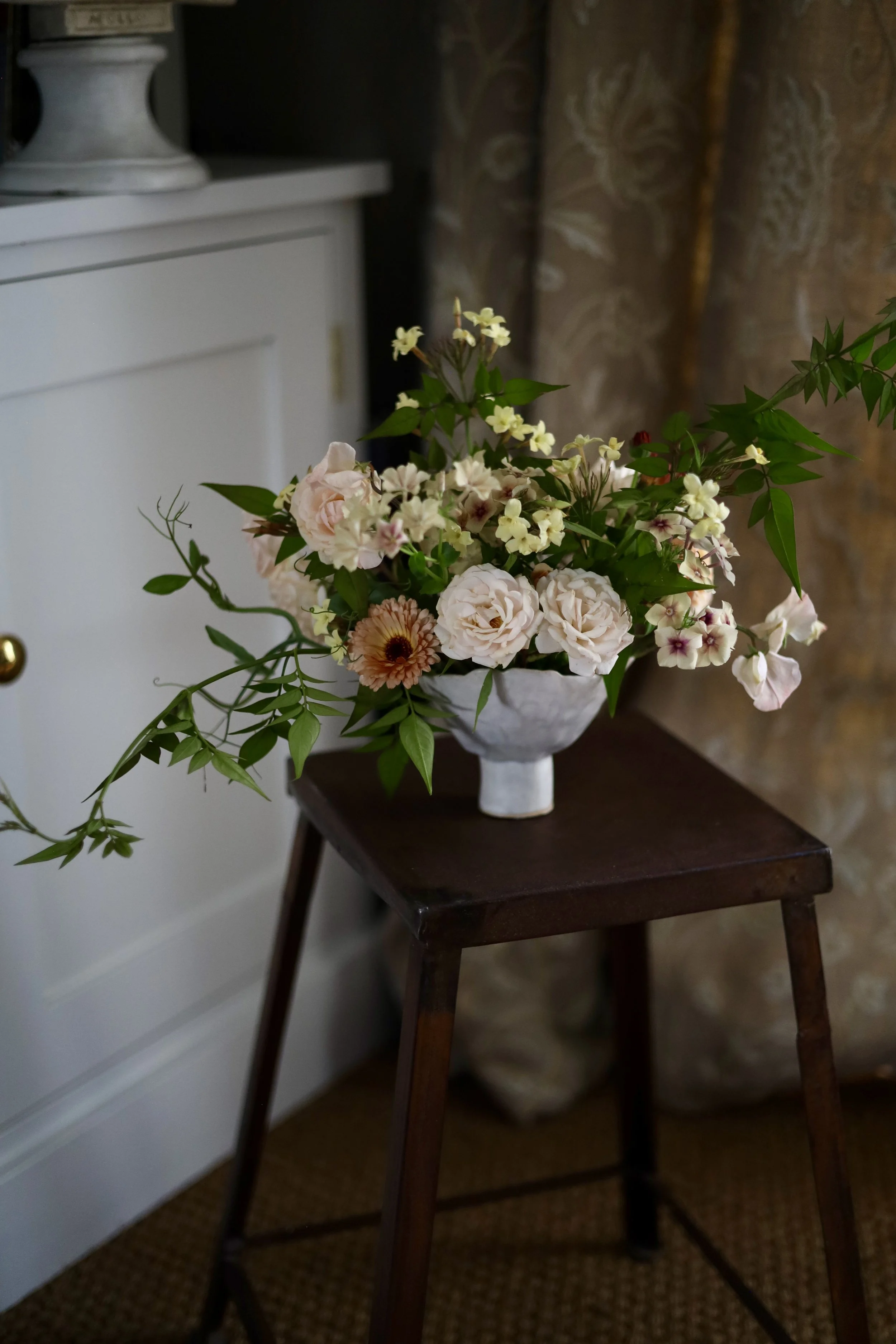 A bouquet of various flowers, including roses and other blooms, arranged in a white vase, placed on a wooden stool in a cozy indoor setting.