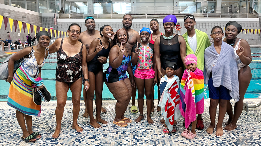 Group of people, including children and adults, at an indoor swimming pool, many wearing swimsuits and towels, smiling and posing for the camera.