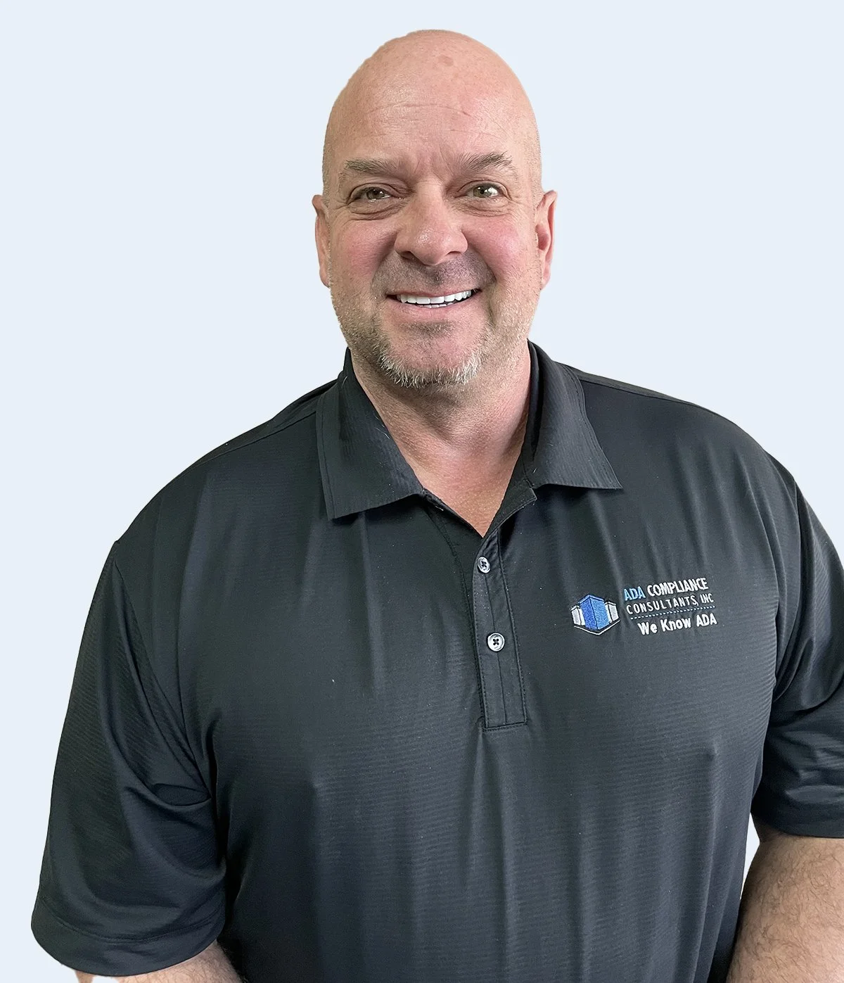 A smiling man with a bald head and a goatee wearing a black polo shirt with a logo and text, standing against a light gray background.