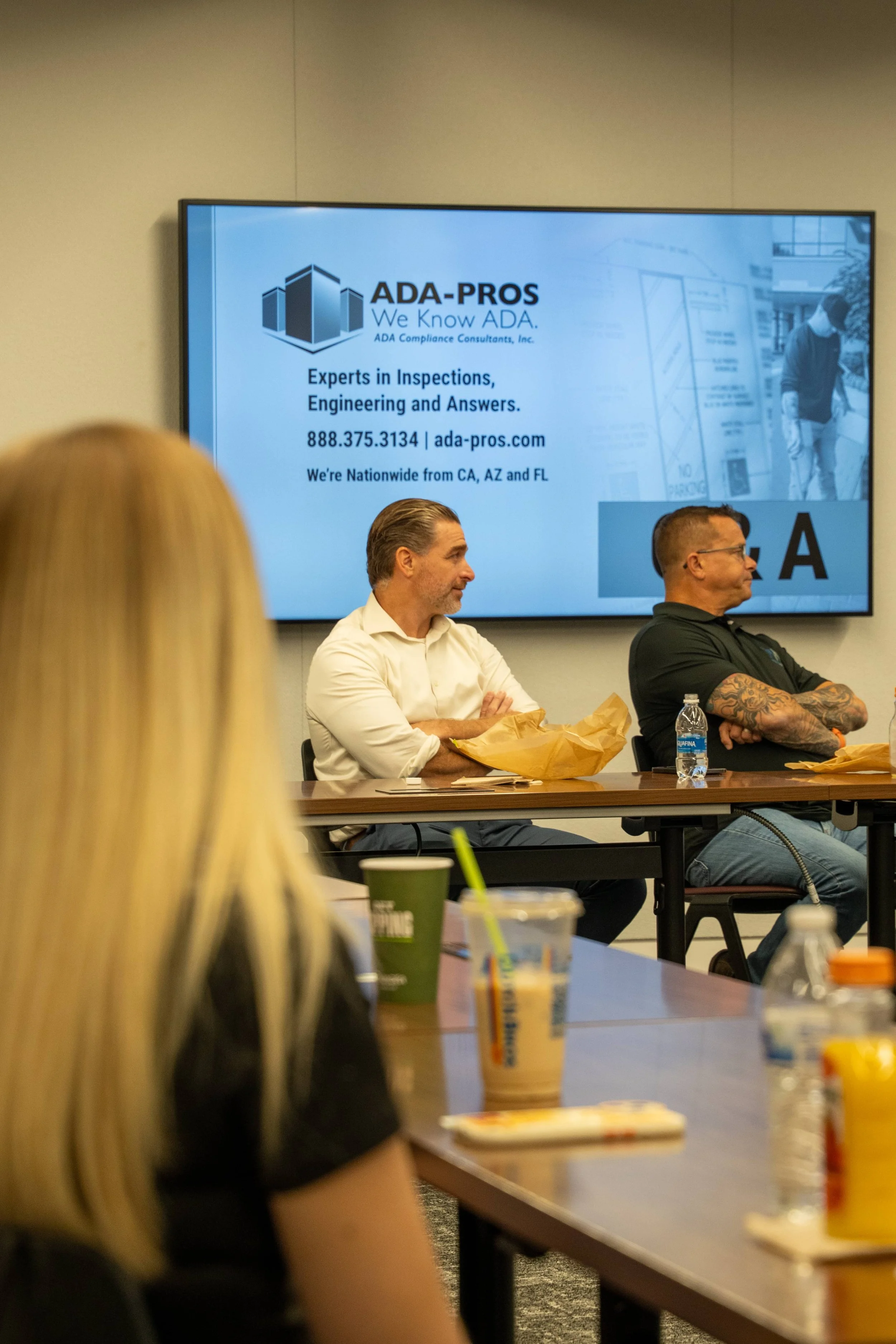 A presentation screen displaying the logo and contact information for ADA-PROS, a company that specializes in inspections and engineering services, with people seated at a table in a meeting room.