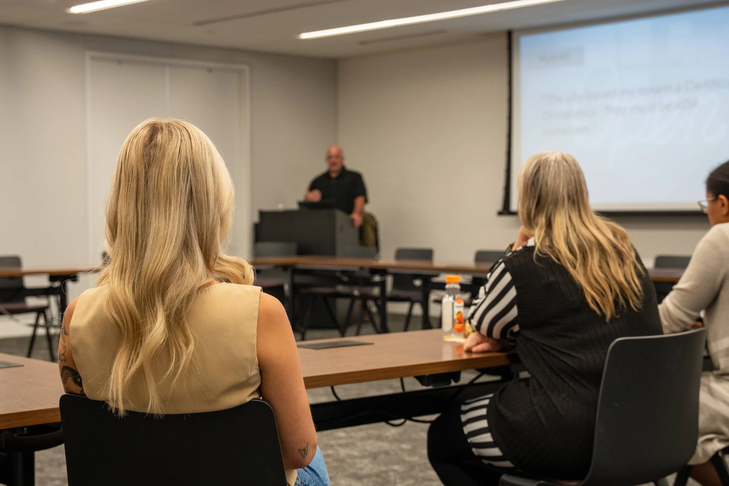 A woman with long blonde hair and a tattoo on her arm is seated at a conference room table, facing a presenter who is standing near a projector screen. Other attendees are also seated at the table, listening to the presentation.