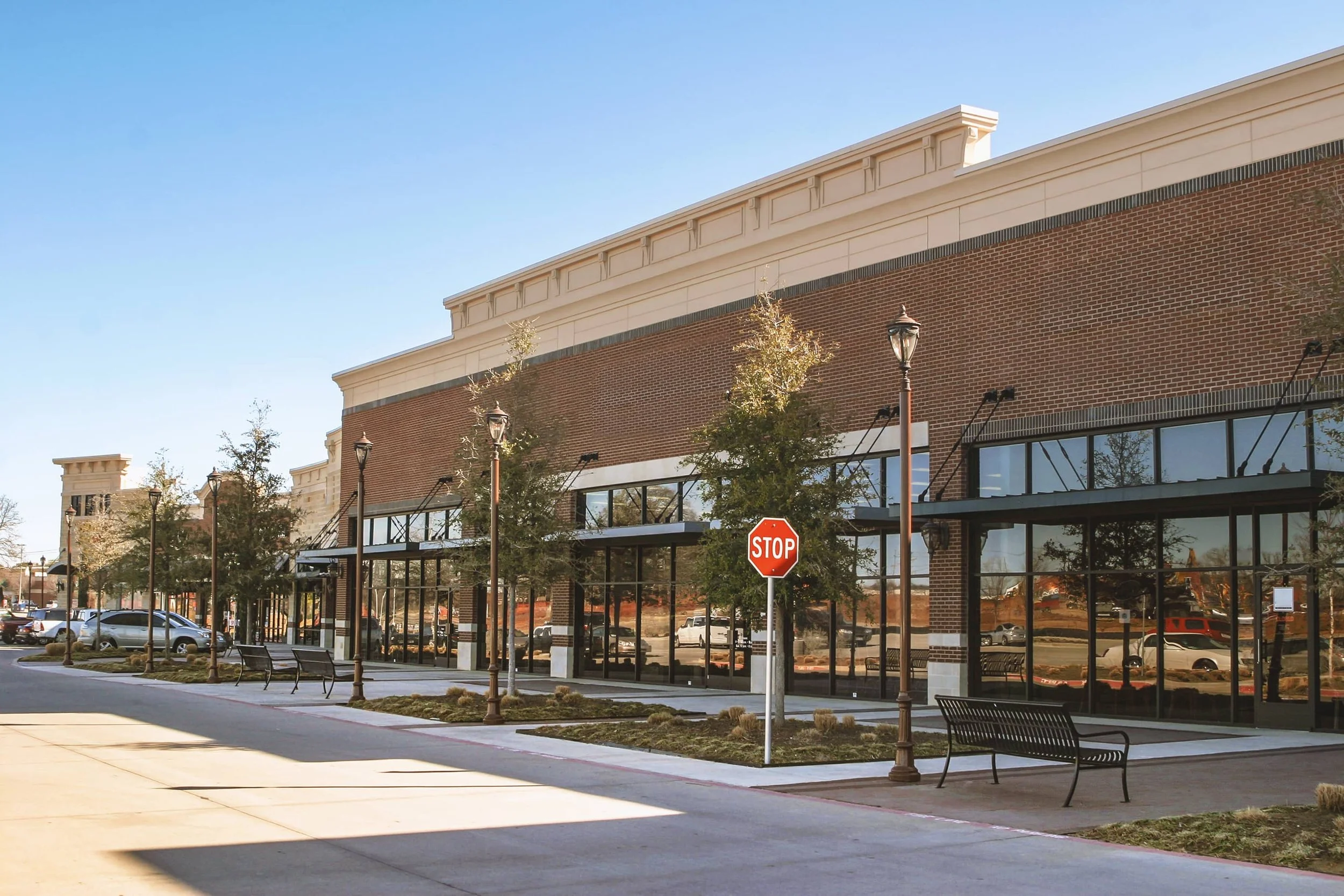 A modern shopping center with brick and glass exterior, parking lot with cars, street lamps, and benches, under a clear blue sky during daytime.