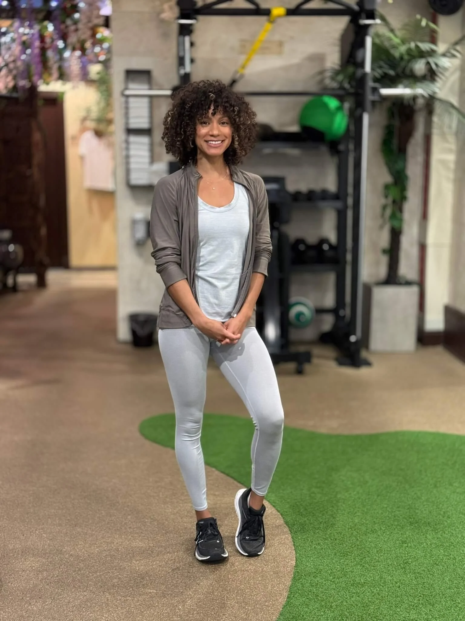 Woman with curly hair smiling in a gym, wearing a light gray top and leggings with a zip-up jacket, standing in front of fitness equipment.