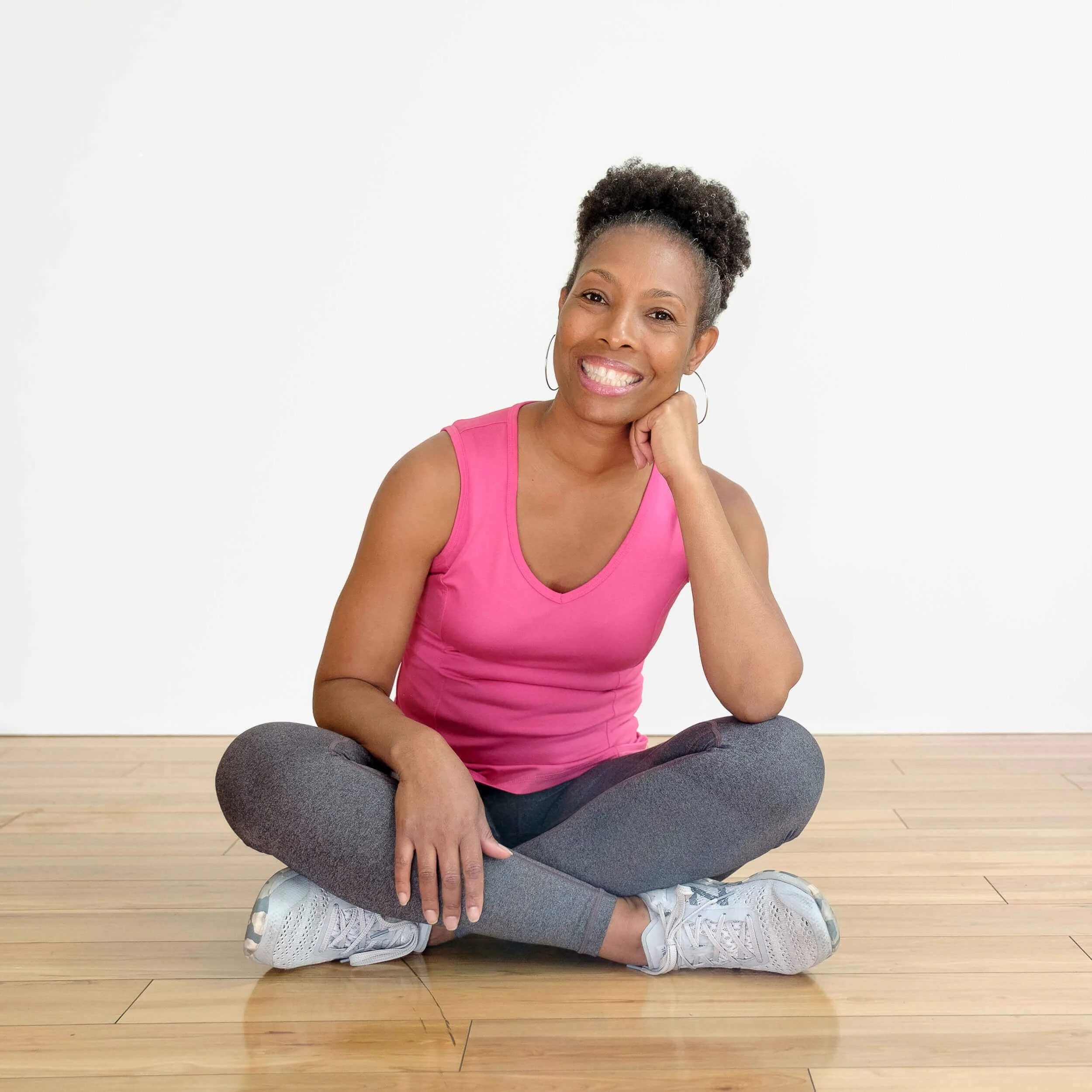 A woman sitting on a wooden floor, smiling, wearing a pink sleeveless top, gray leggings, white sneakers, with short curly hair, and hoop earrings, against a plain white wall.