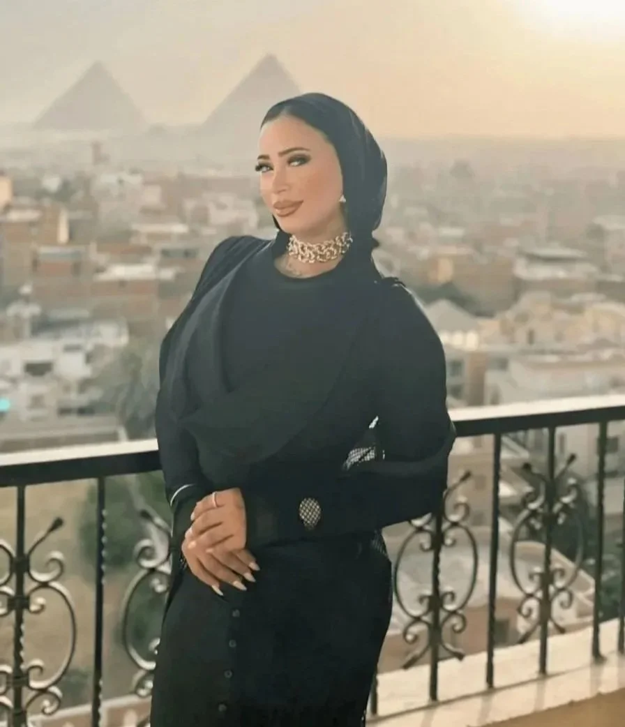 A woman standing on a balcony with a cityscape in the background, including the pyramids of Egypt visible in the distance, during sunset.