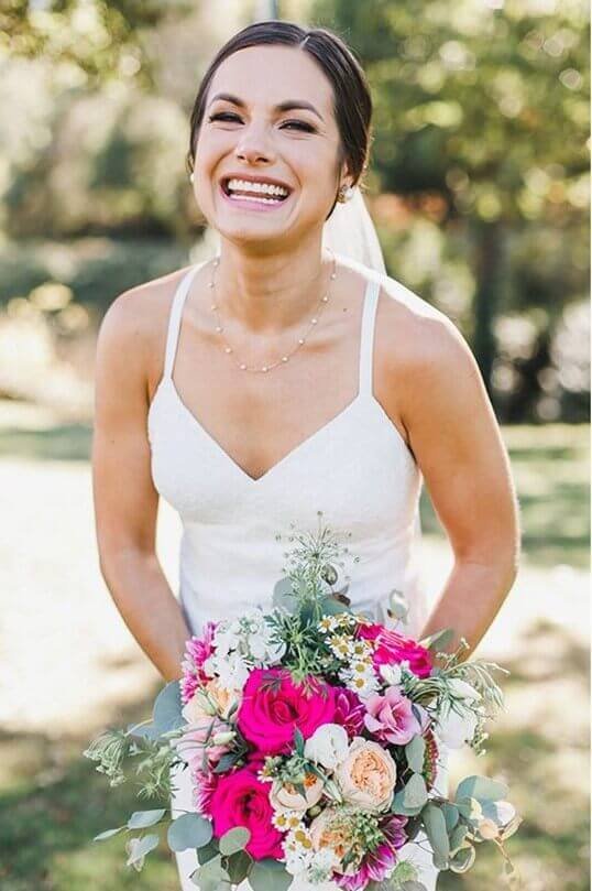Smiling bride in a white wedding dress holding a colorful bouquet outdoors on her wedding day.