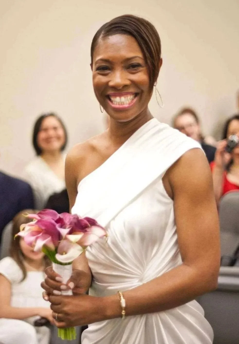 Smiling bride in a white one-shoulder wedding dress holding a bouquet during a ceremony with guests in the background.