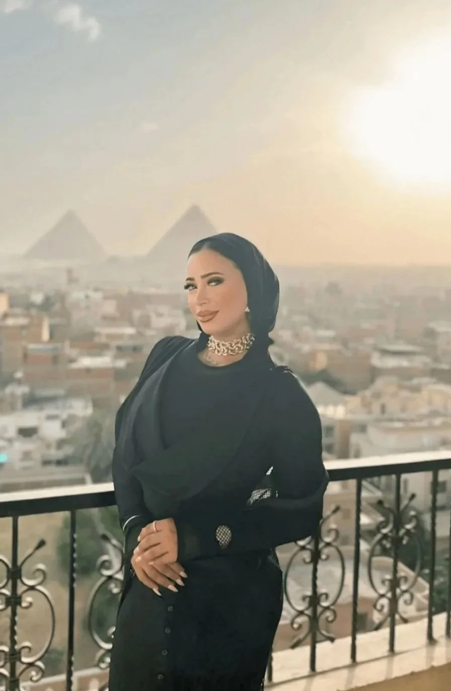 A woman standing on a balcony with a cityscape in the background, including the pyramids of Egypt visible in the distance, during sunset.