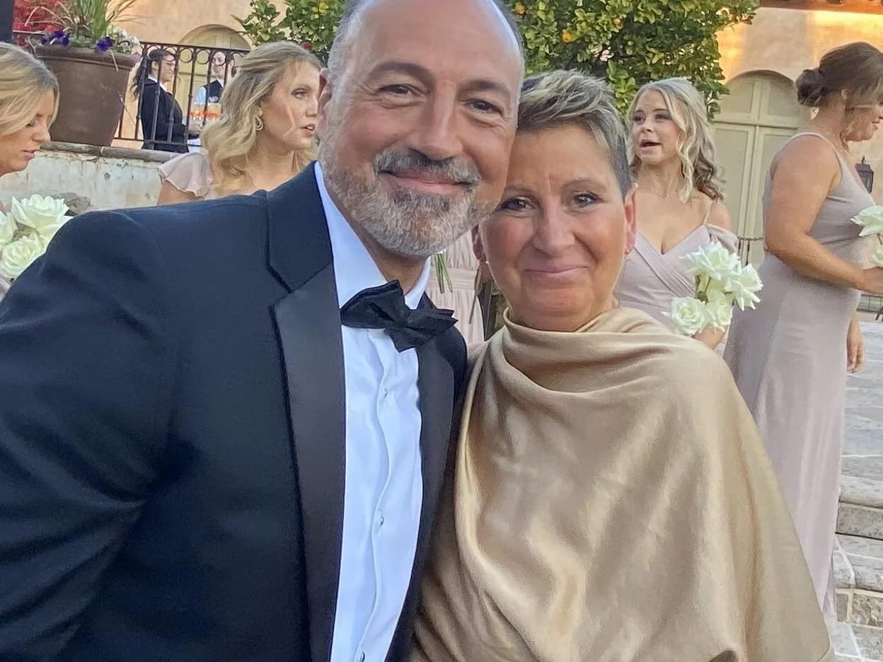 Smiling mom-of-a-bride wearing a gold dress standing with her husband wearing a black tuxedo