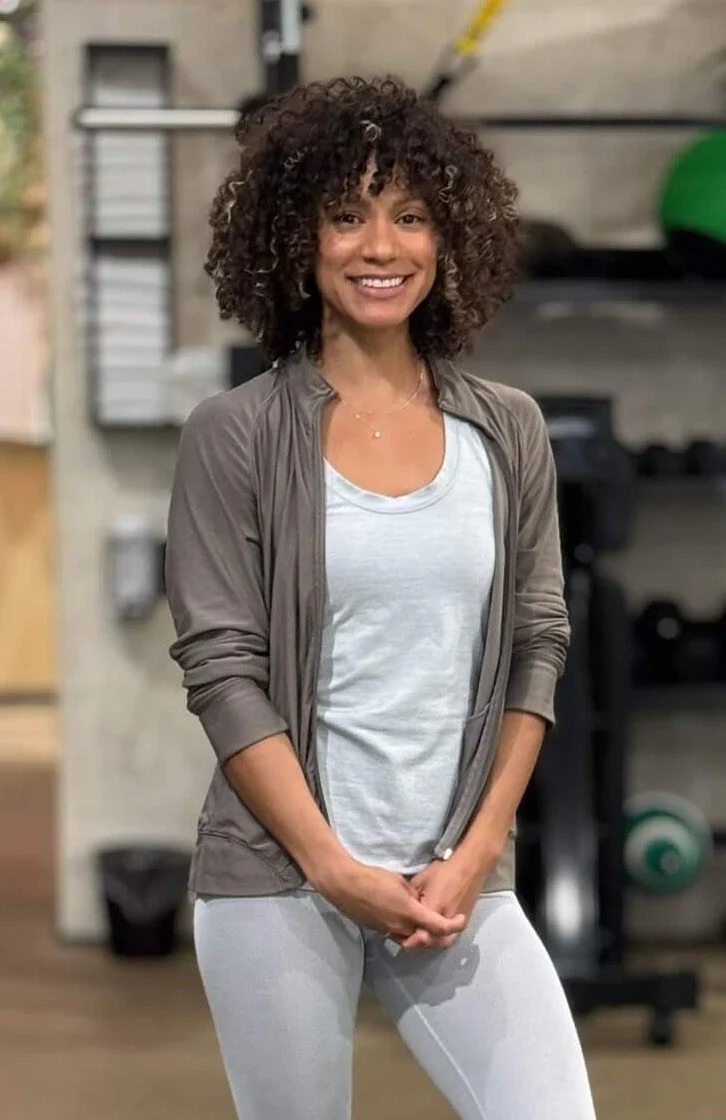 Woman with curly hair smiling in a gym, wearing a light gray top and leggings with a zip-up jacket, standing in front of fitness equipment.