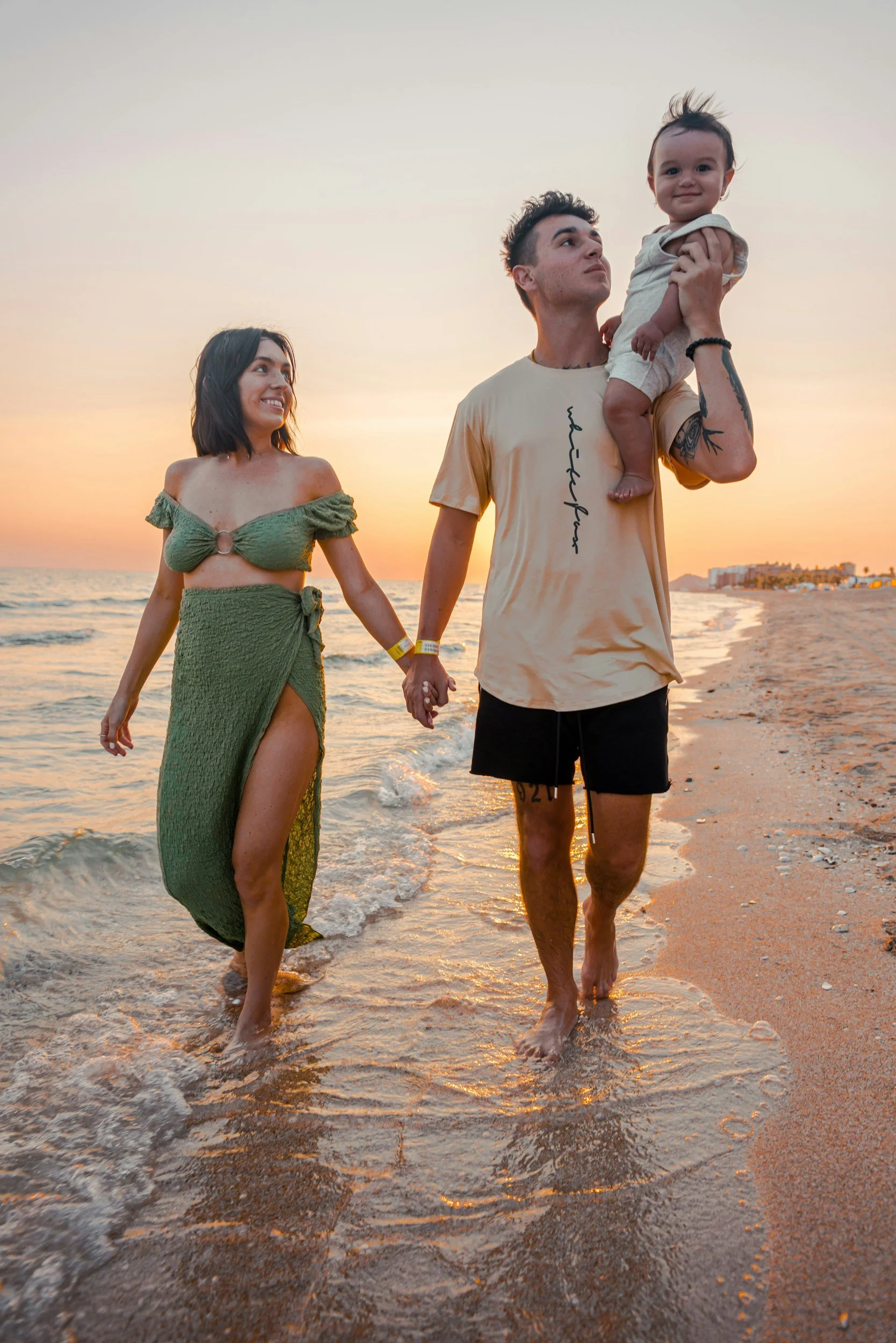 A family of three walking along the beach at sunset. The mother is wearing a green two-piece dress, the father in a beige T-shirt and black shorts, holding a young girl in his arm. They are holding hands and smiling, with the ocean and a sunset in the background.