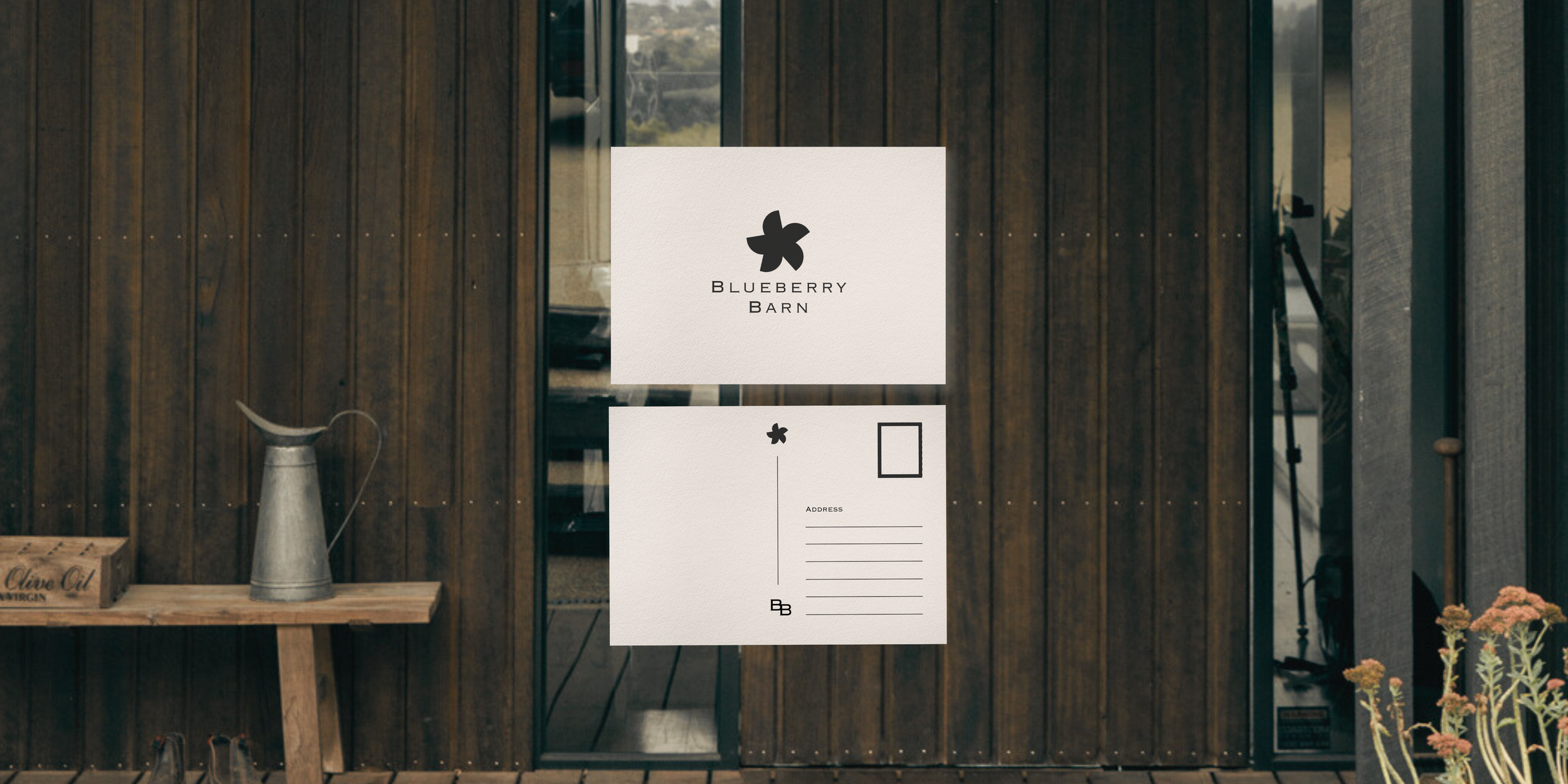 Wooden exterior wall with a door. In front, there's a sign for Blueberry Barn with a stylized blueberry logo. Below it, there is a smaller sign with an address section and space for contact information. A watering can is placed on a wooden table to t